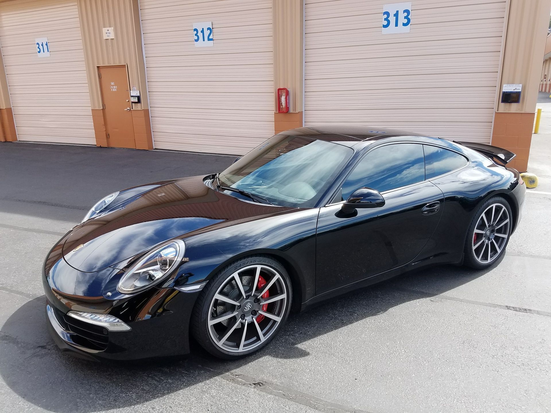 Black Porsche 911 sports car parked outside storage units with doors numbered 312 and 313.