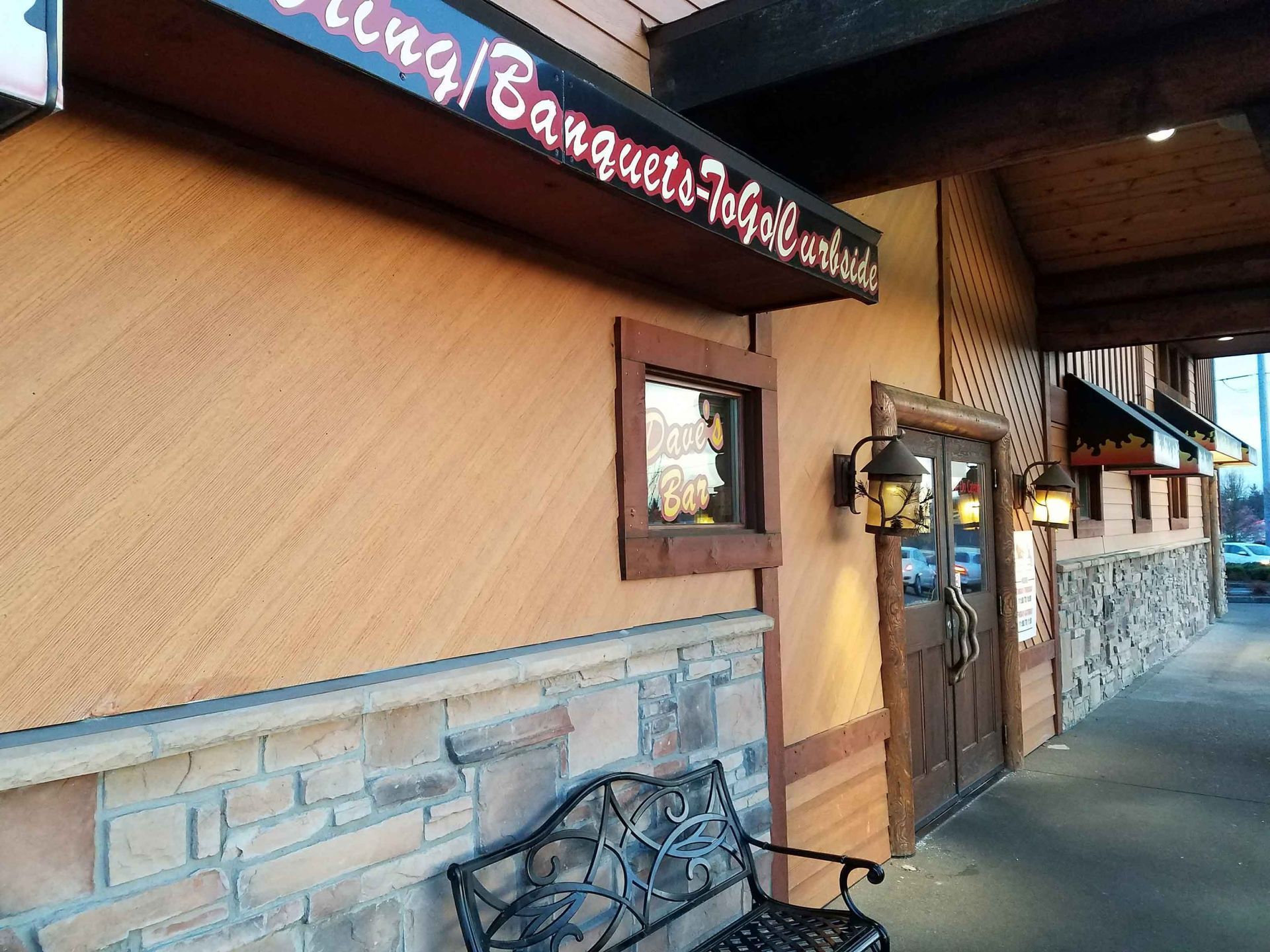 Restaurant exterior with tan stucco, stone accents, and a wooden door. A bench sits below a sign for