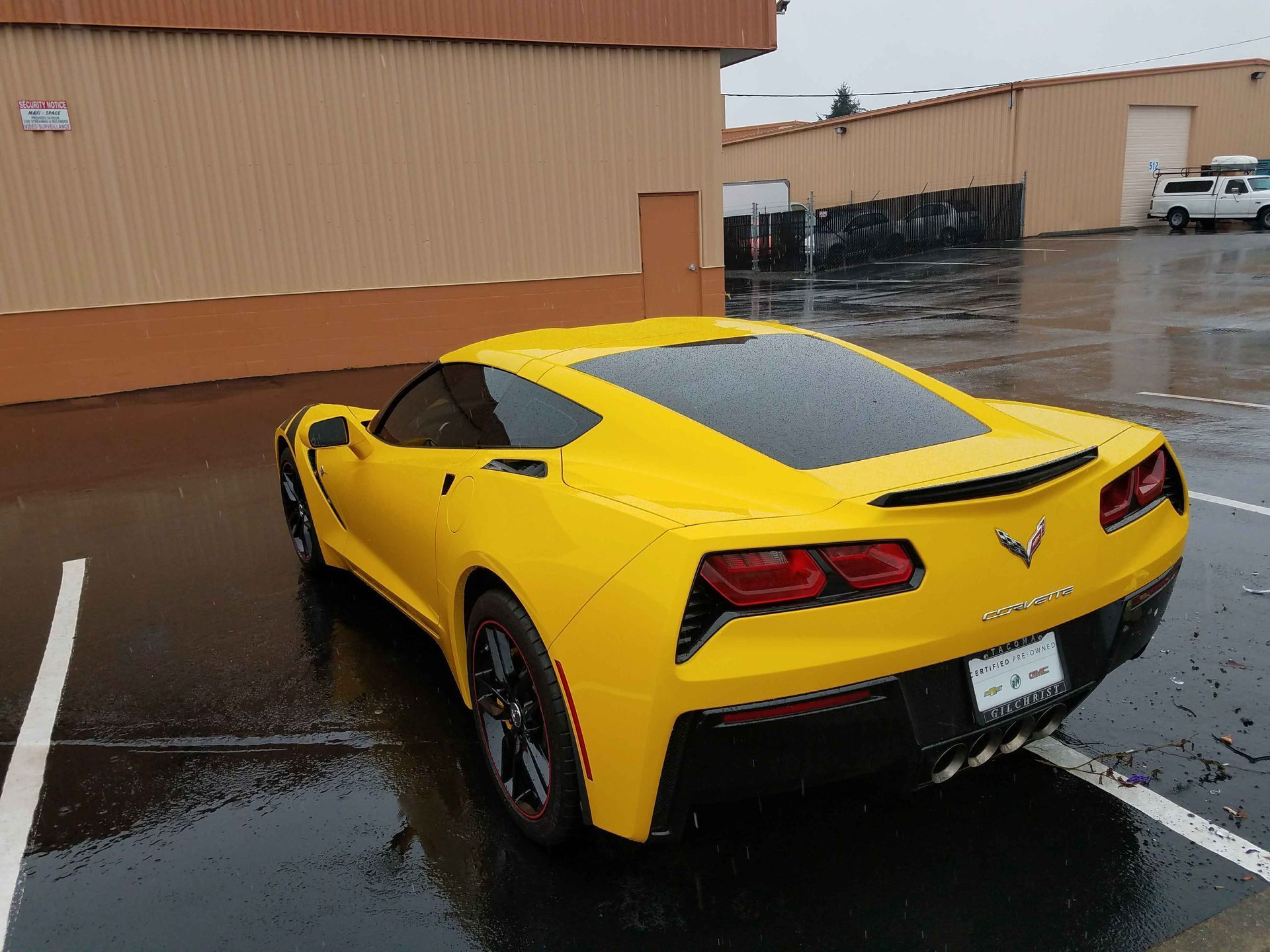 Yellow Corvette sports car parked in a wet lot, with tan buildings in the background.