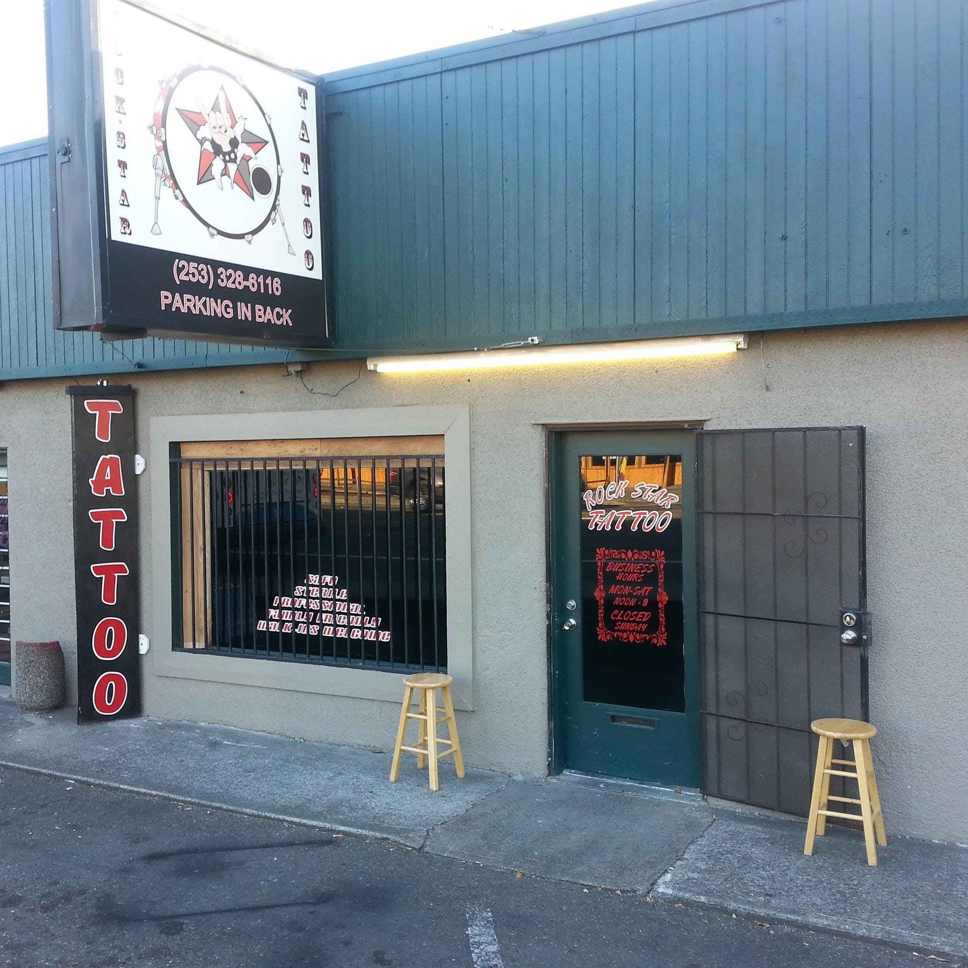 Exterior of a tattoo parlor; sign with a star logo; door open; two stools outside.