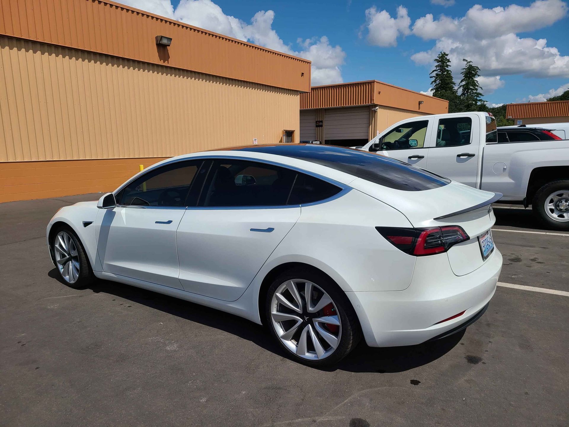 White Tesla sedan parked next to a white pickup truck in a parking lot with a building in the background.