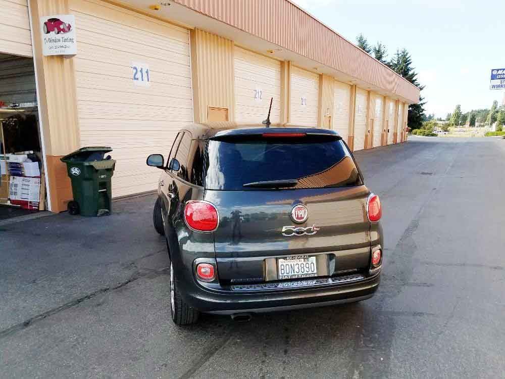 Dark gray Fiat 500L parked outside beige storage units. Asphalt driveway, green trash can, sunny day.