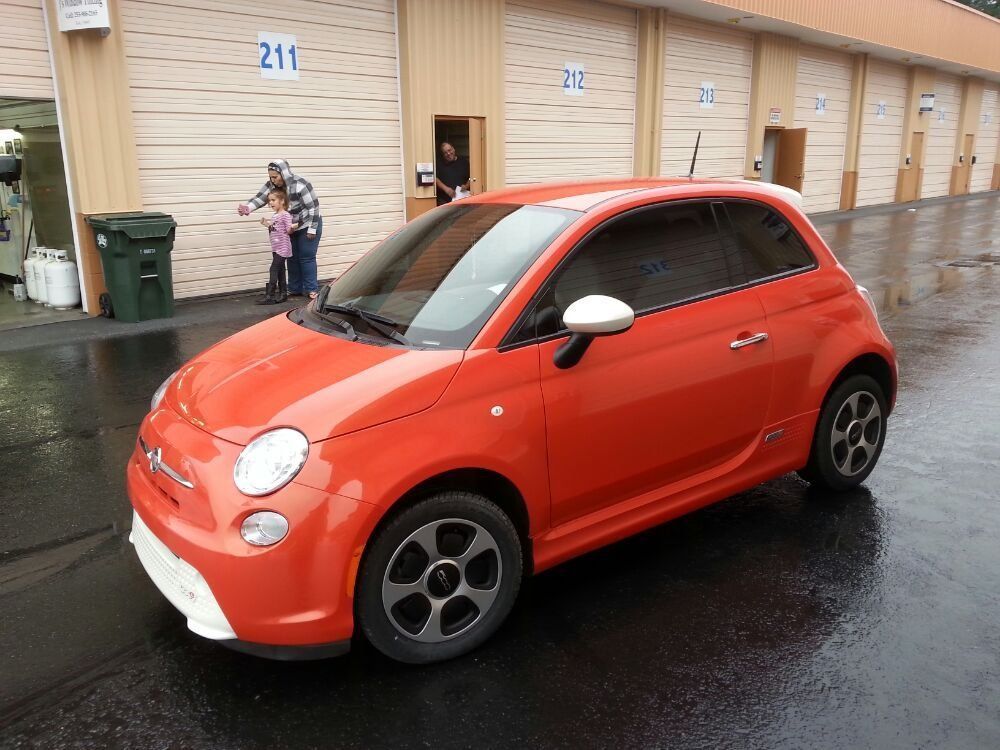 Orange Fiat 500e electric car parked near storage units, with people near door 211.
