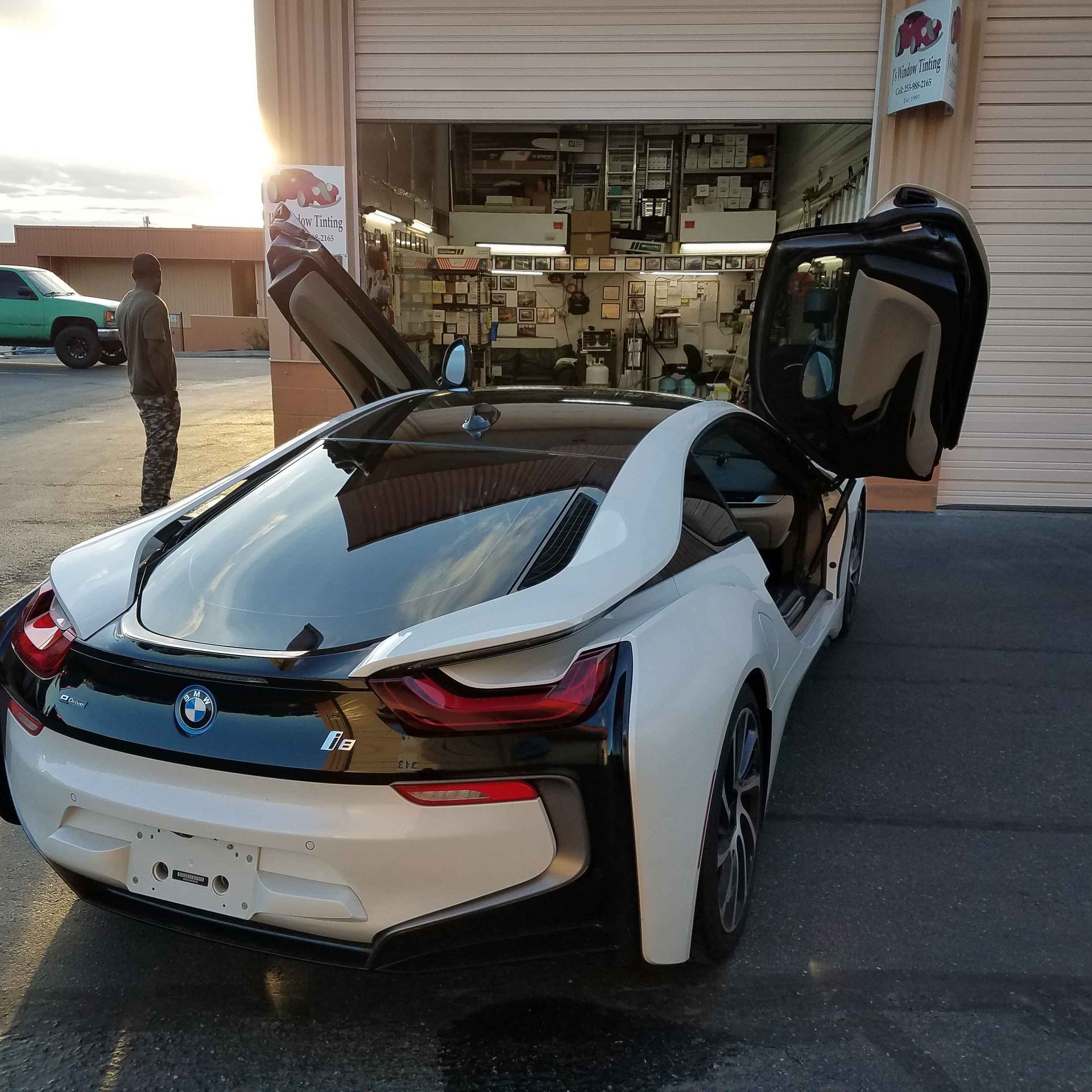 White BMW i8 with doors open, parked near a garage, person standing in the background.