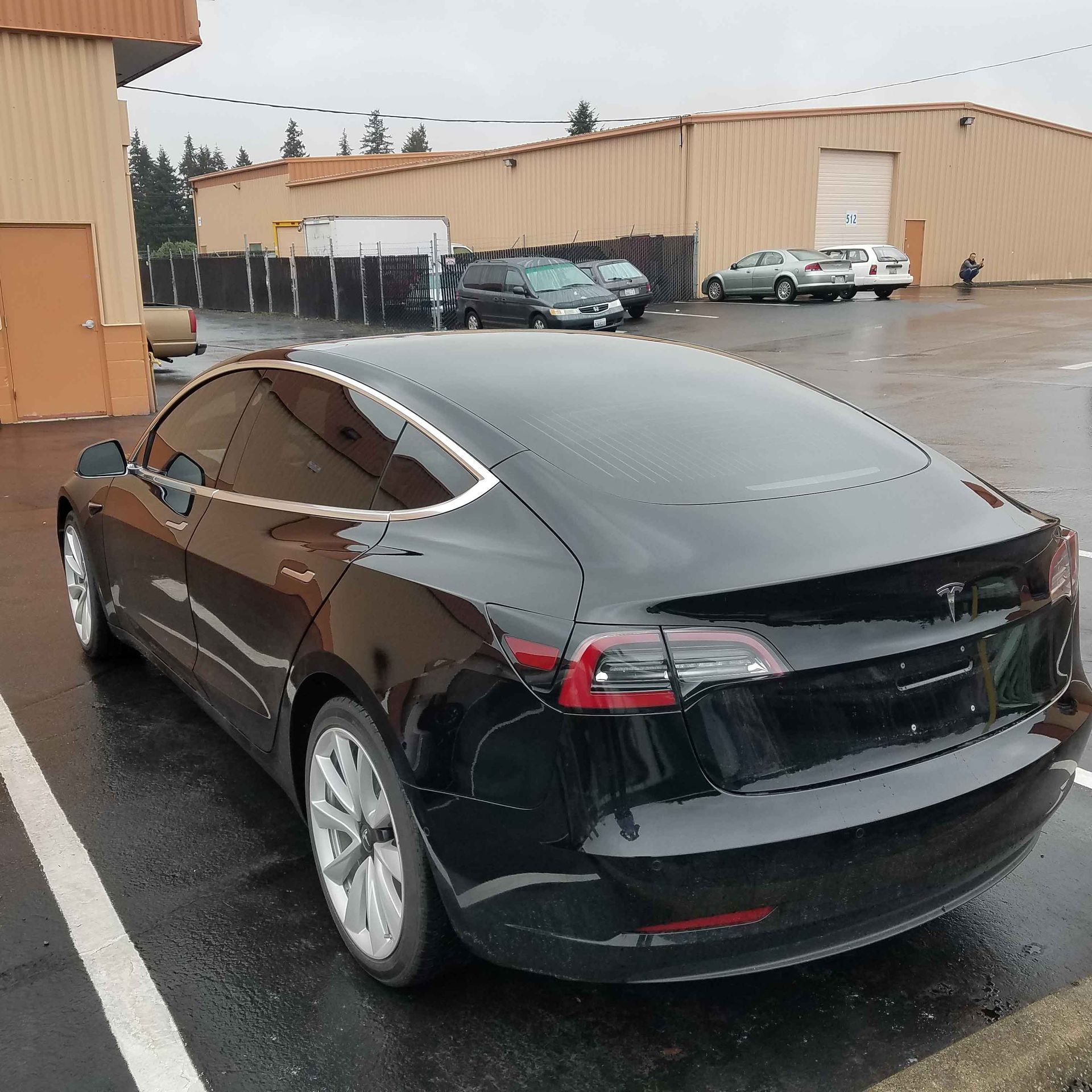 Black Tesla Model 3 parked in a wet parking lot in front of industrial buildings.