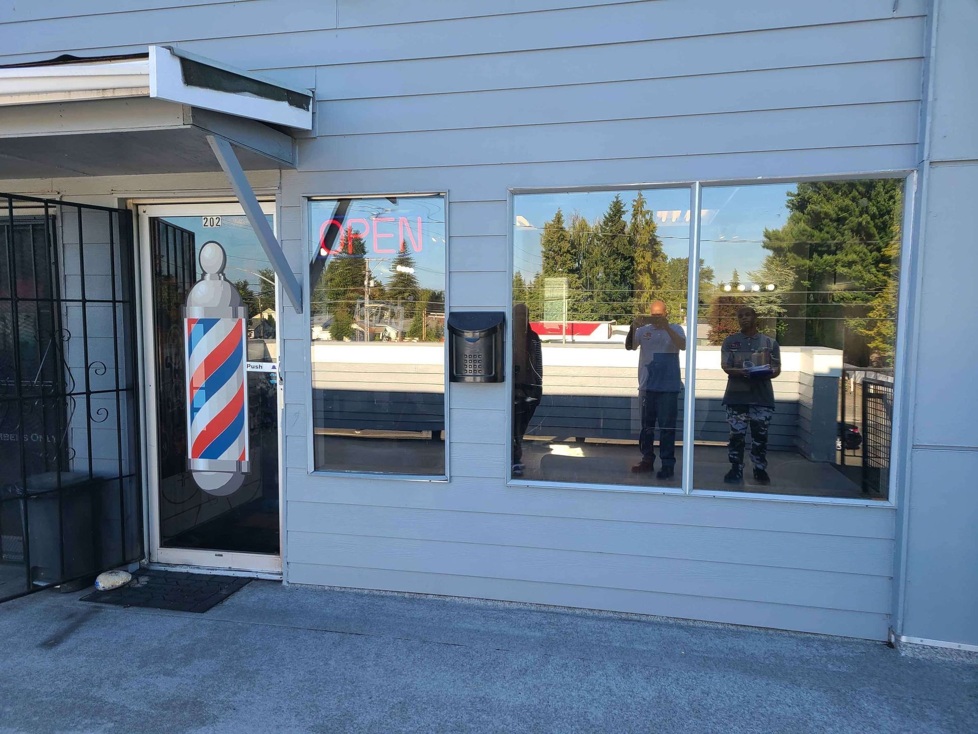 Exterior of a barbershop. Blue building, barber pole, two people reflected in the windows.