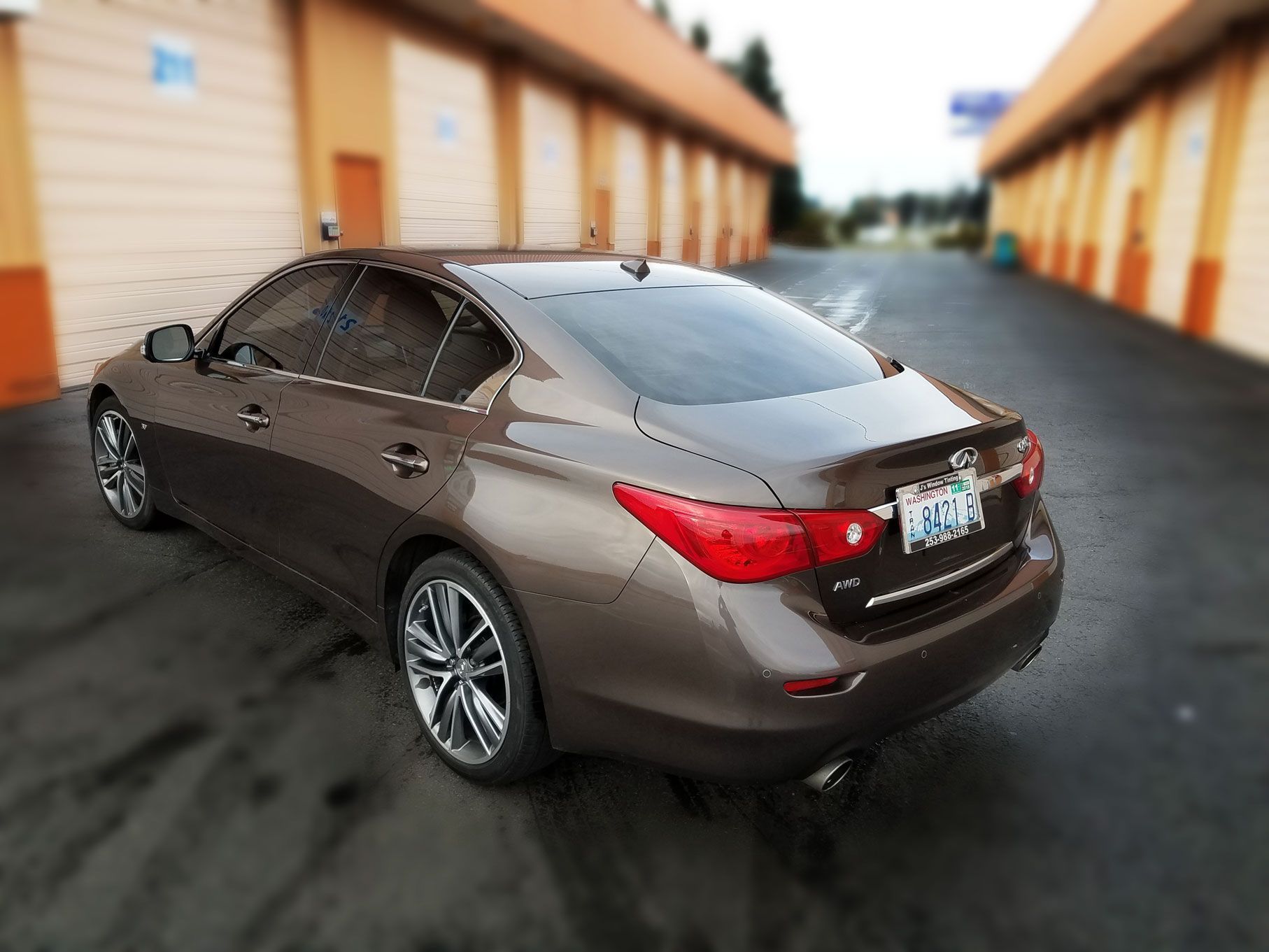 Brown Infiniti sedan parked on wet pavement near storage units.