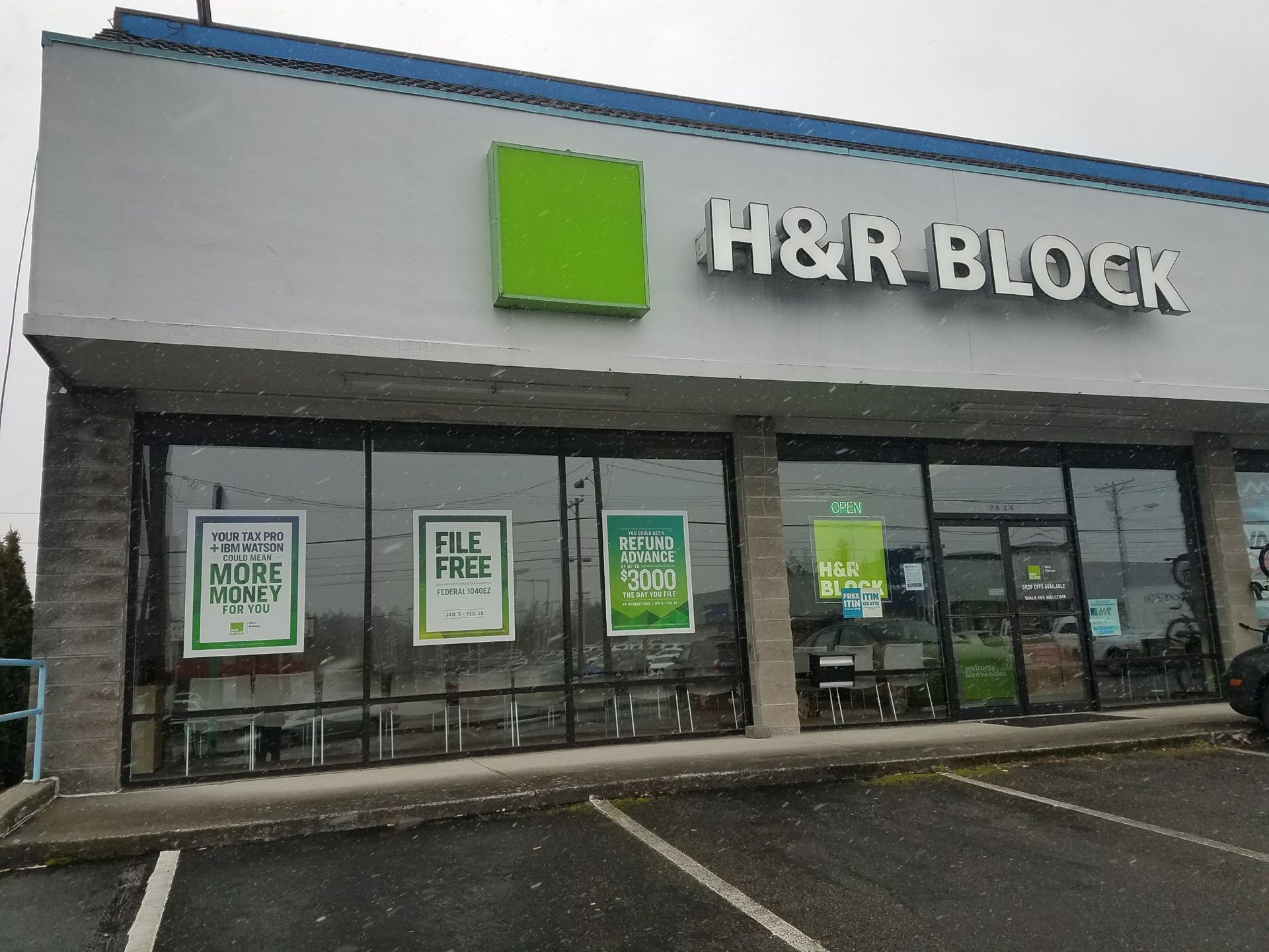 H&R Block storefront with green and white signs. Grey building, overcast sky, parking lot.