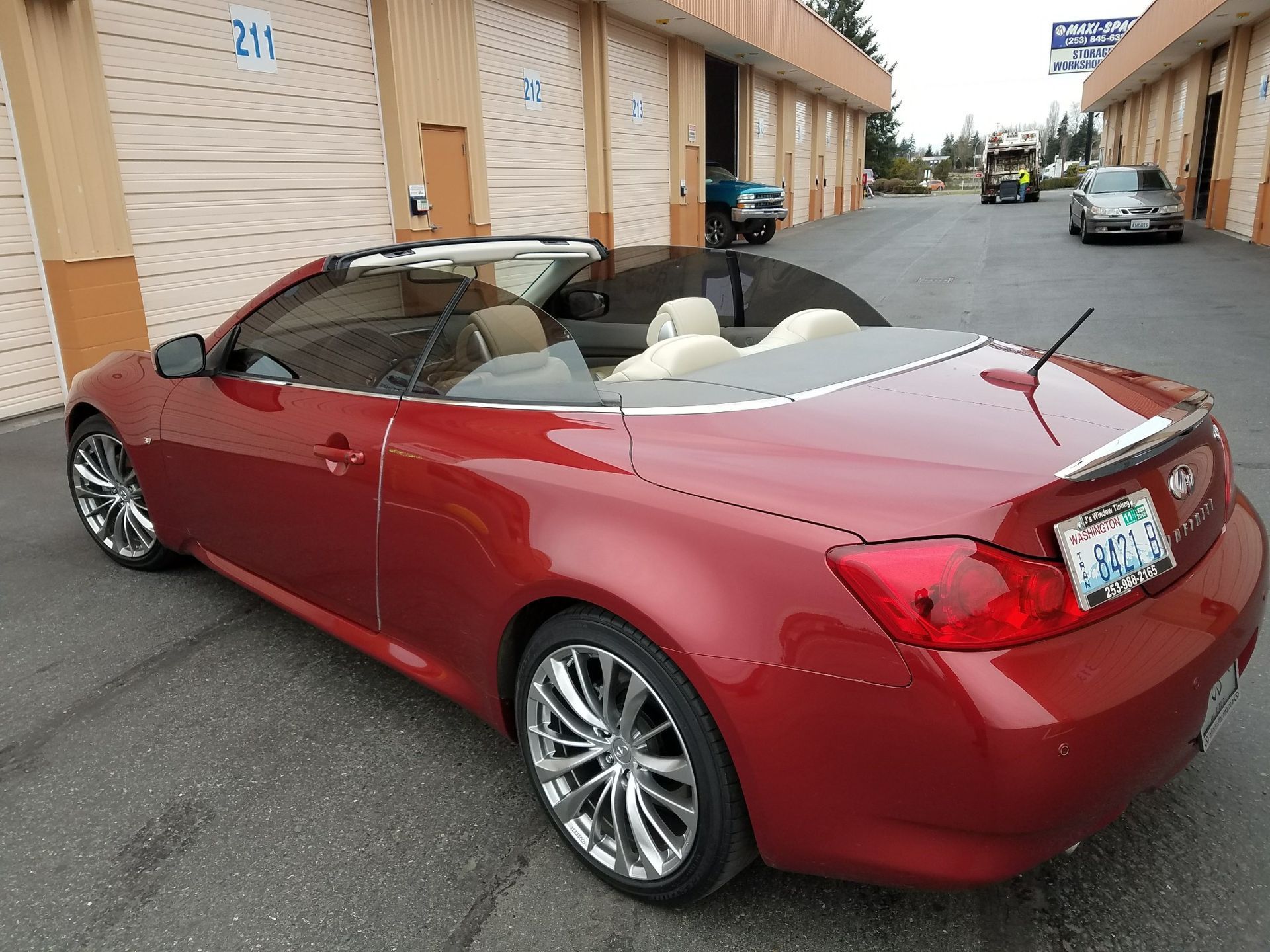 Red convertible car parked in front of storage units. Chrome wheels, white interior, top down.