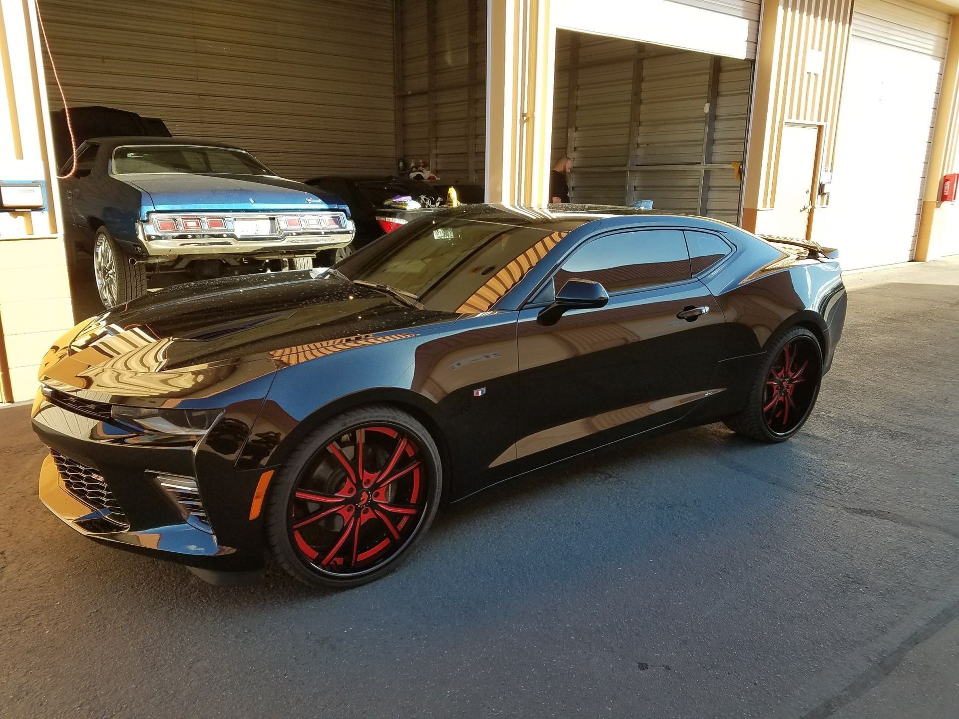 Black Camaro with red rims parked outside a garage, another car in the background.