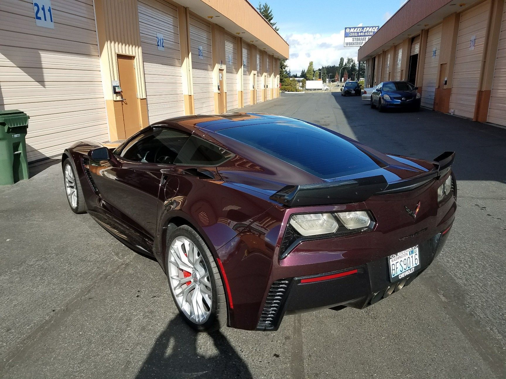 Maroon Corvette parked on a paved area in front of commercial buildings on a sunny day.