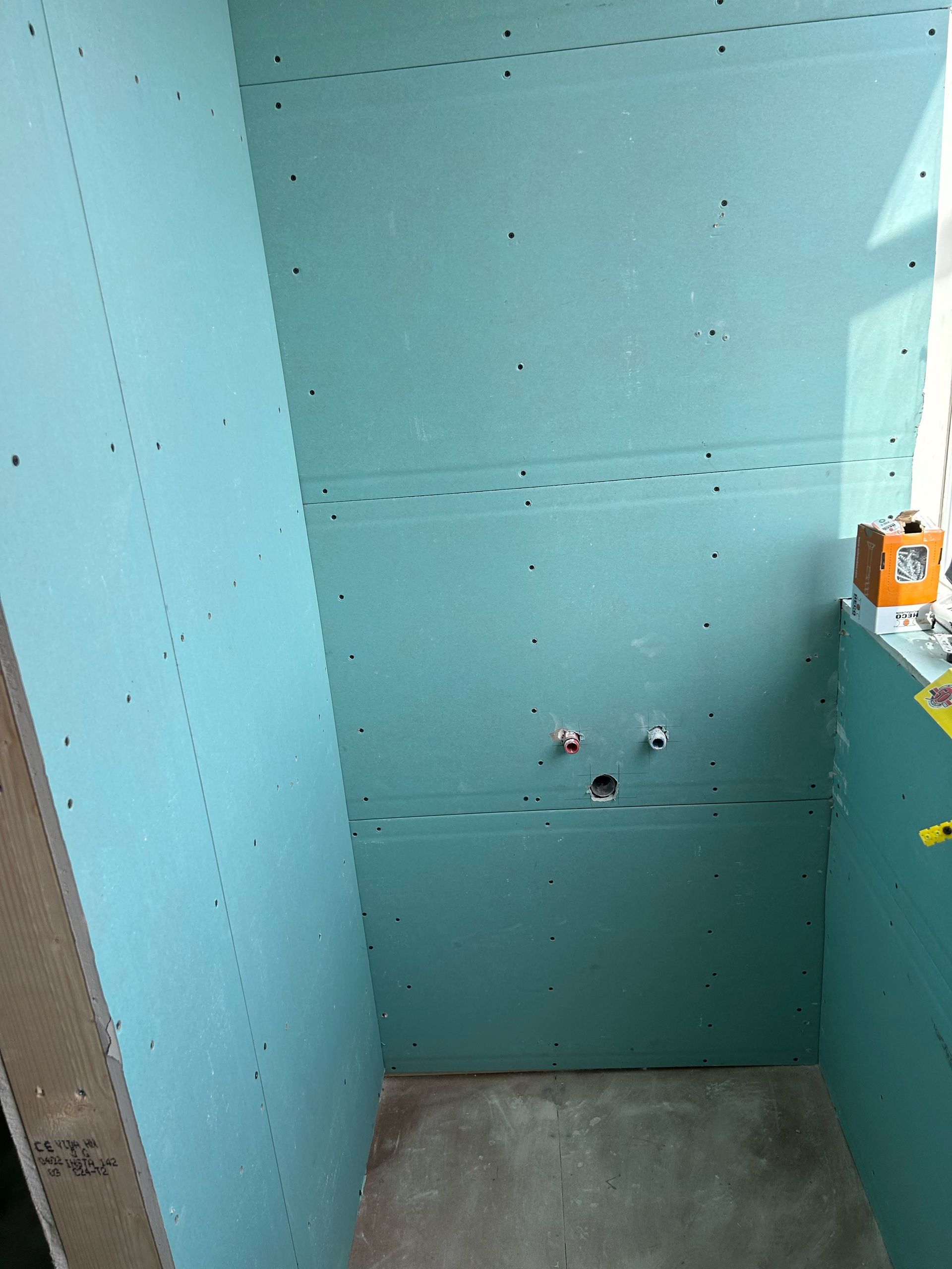 Een douchecabine in aanbouw met blauwe vochtwerende gipsplaten, zichtbare leidingen en een betonnen vloer.