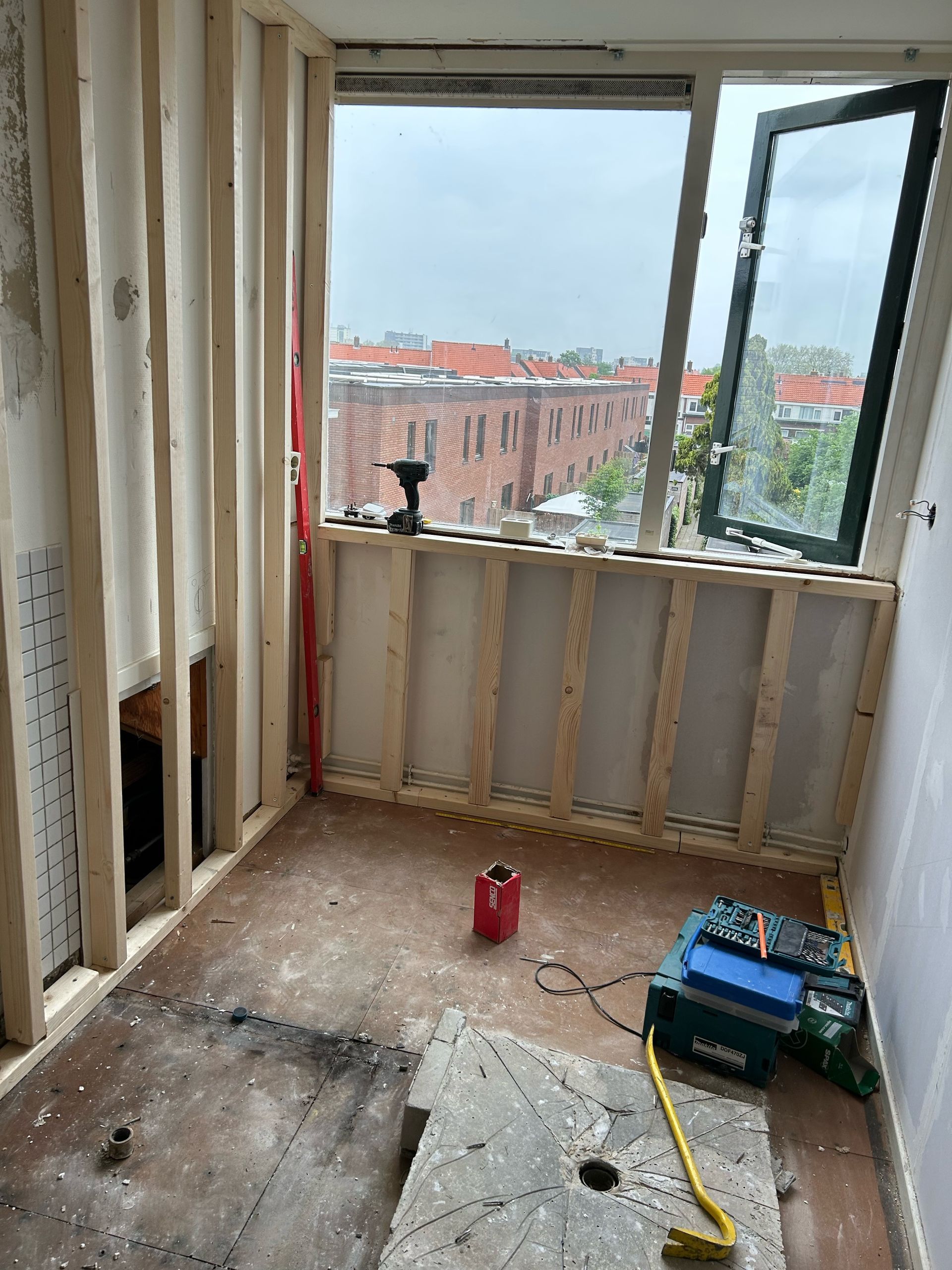 Een ruimte in aanbouw met houten wandconstructie, zichtbare vloer, een groot raam en gereedschap verspreid over de vloer.