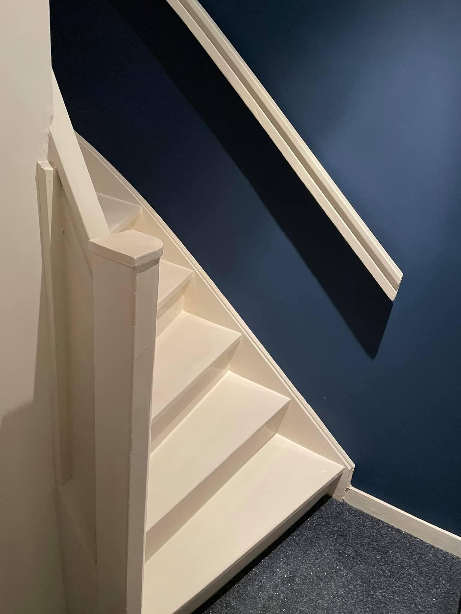 Een lichtgekleurde houten trap met een witte leuning tegen een donkerblauwe muur.