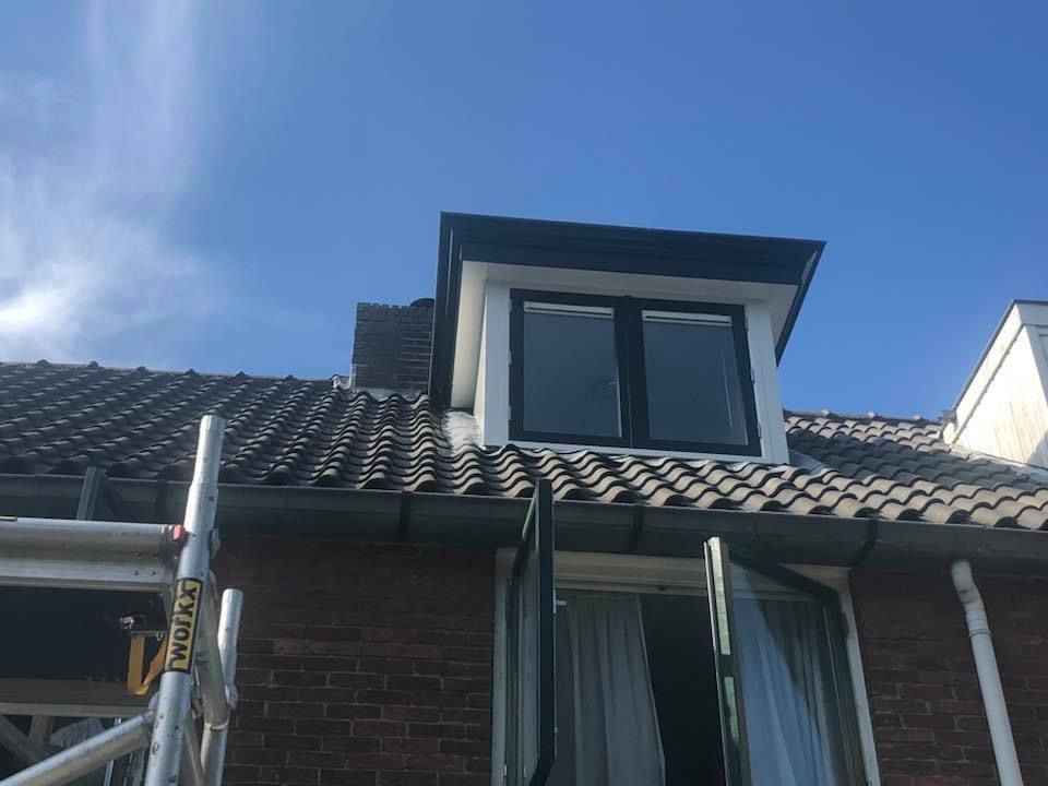 Een close-up van een bakstenen dak met een dakkapel met zwarte kozijnen en een stuk steigerwerk in de buurt.