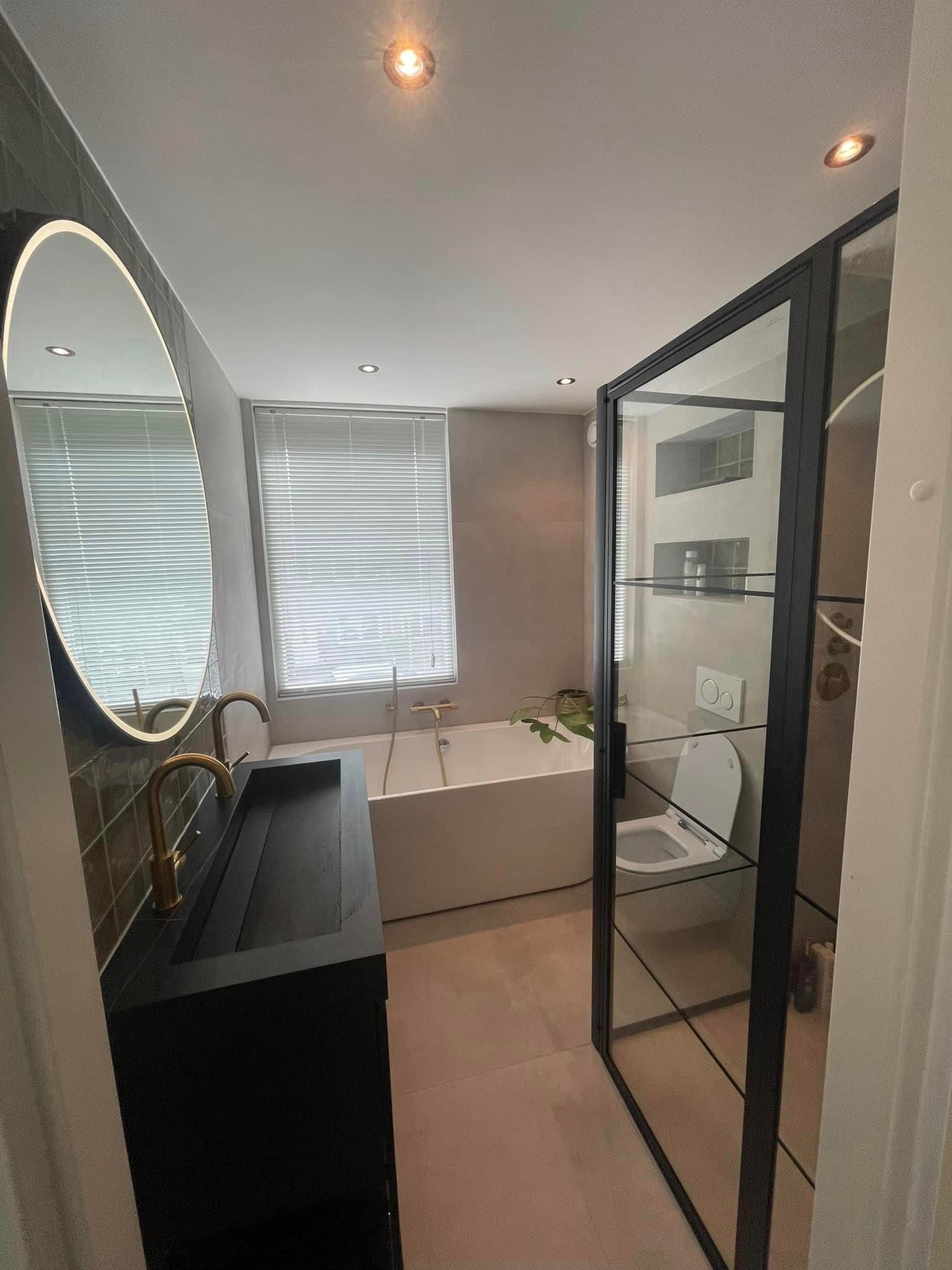 Een moderne badkamer is voorzien van een donker badmeubel met een ronde spiegel met achtergrondverlichting, een toilet met glazen cabine en een wit bad.