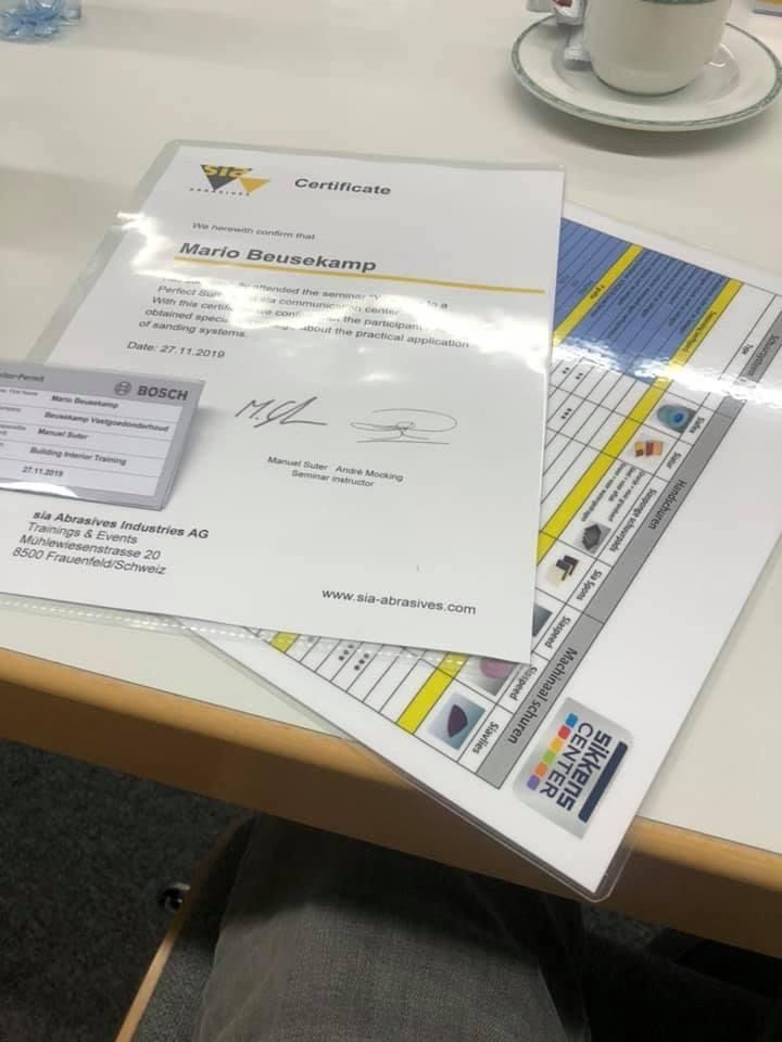 Een certificaat voor Mario Braukamp, een klein naamplaatje en een overzichtstabel liggen op een witte tafel naast een kopje thee.