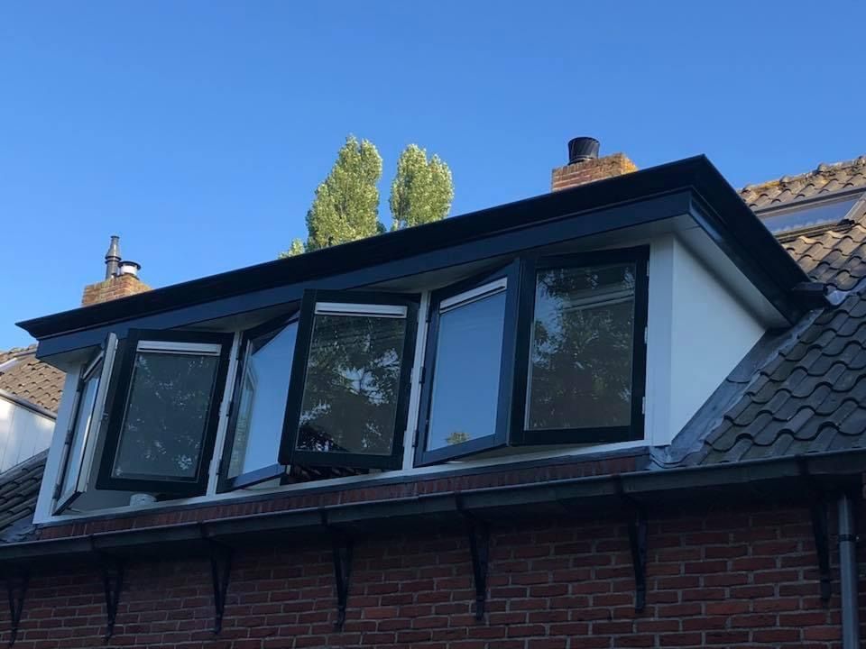 Een dakkapel met zwarte kozijnen, geplaatst op een rood bakstenen dak, tegen een helderblauwe hemel.