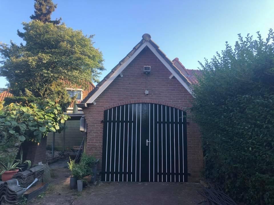 Een stenen garage met een puntdak en grote, zwart-wit gestreepte deuren, gelegen in een tuin met bomen en struiken.