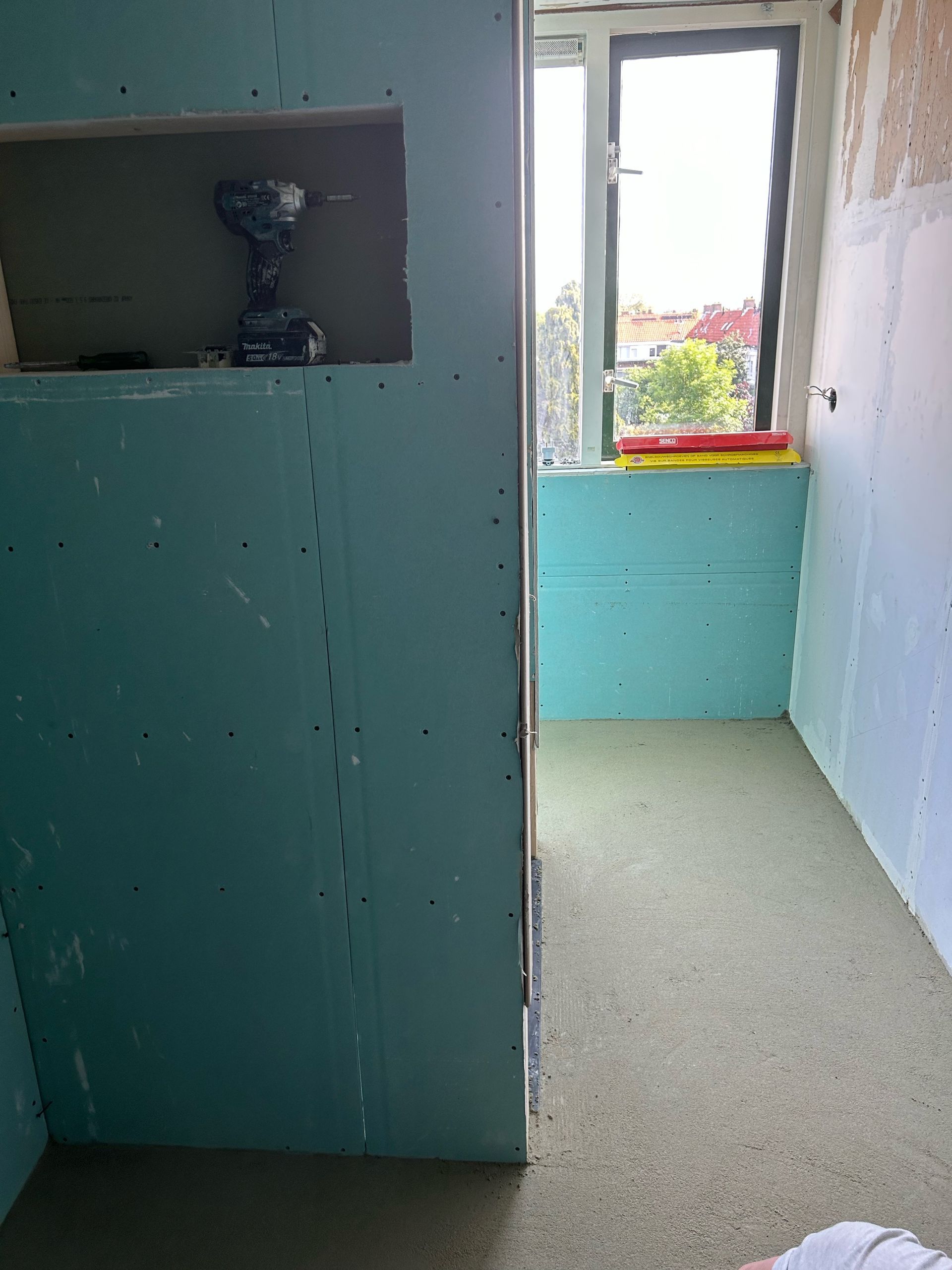 Een kamer in aanbouw met turquoise gipsplaten, een raam en een accuboormachine in een uitsparing in de muur.