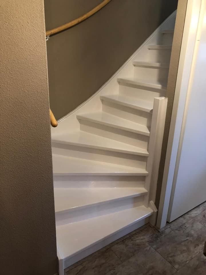 Een witte, kronkelende houten trap die naar boven leidt, gezien vanaf de begane grond naast een donkere muur en tegelvloer.