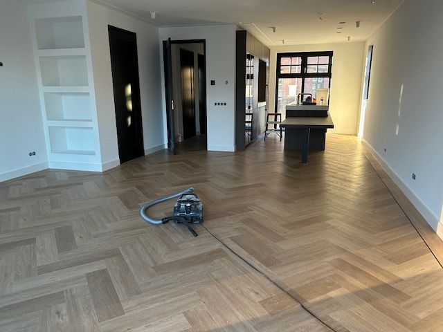 Een lege kamer met een lichte houten vloer in visgraatpatroon, witte muren, ingebouwde planken, donkere deuren en een stofzuiger.