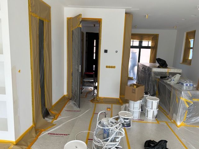 Een kamer die gerenoveerd wordt, met afgeplakte deuropeningen, afgedekte oppervlakken en verfspullen verspreid over een beschermende vloer.