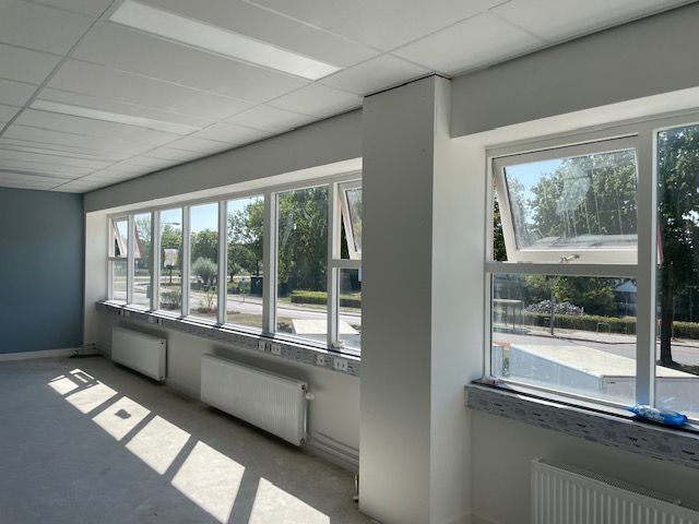 Leeg kantoorinterieur met grote ramen, witte muren, grijze vloer en witte radiatoren onder de ramen.