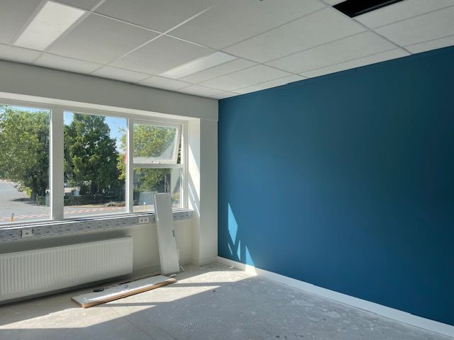 Een leeg kantoor met een groot raam, een radiator en een effen diepblauwe accentmuur onder een wit plafond met tegelpatroon.