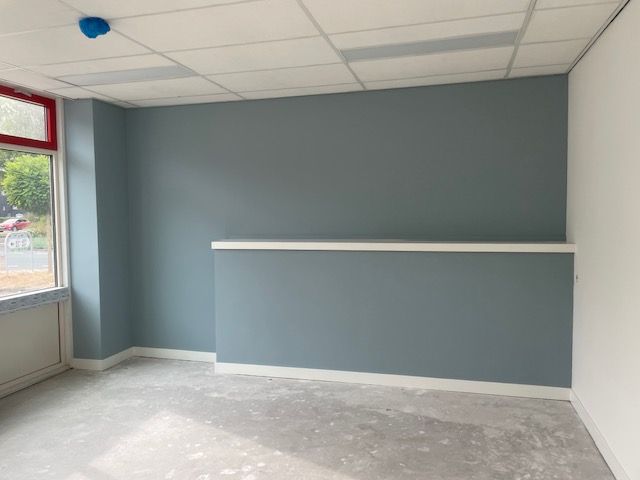 Een lege kamer met blauwgrijze muren, een gedeeltelijke scheidingswand, een wit raamkozijn en een wit systeemplafond.