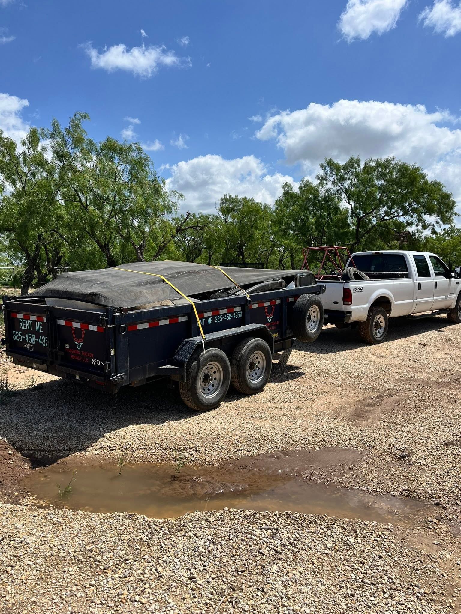 Black utility trailer hitched to a white pickup on a gravel lot with trees and blue sky.