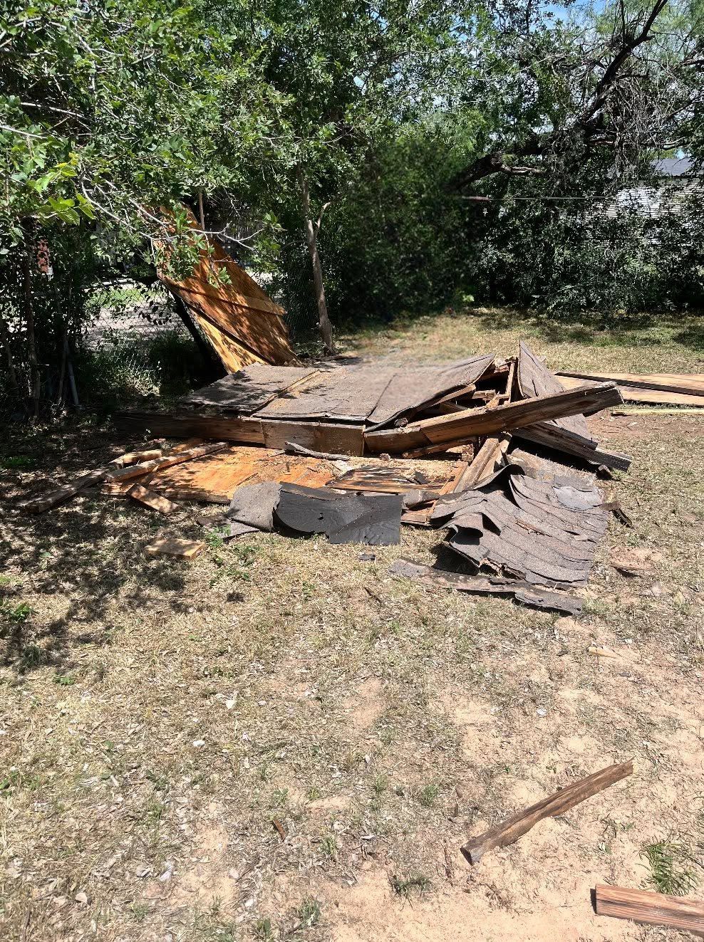 Collapsed wooden structure in a wooded yard with debris and charred remains on the ground