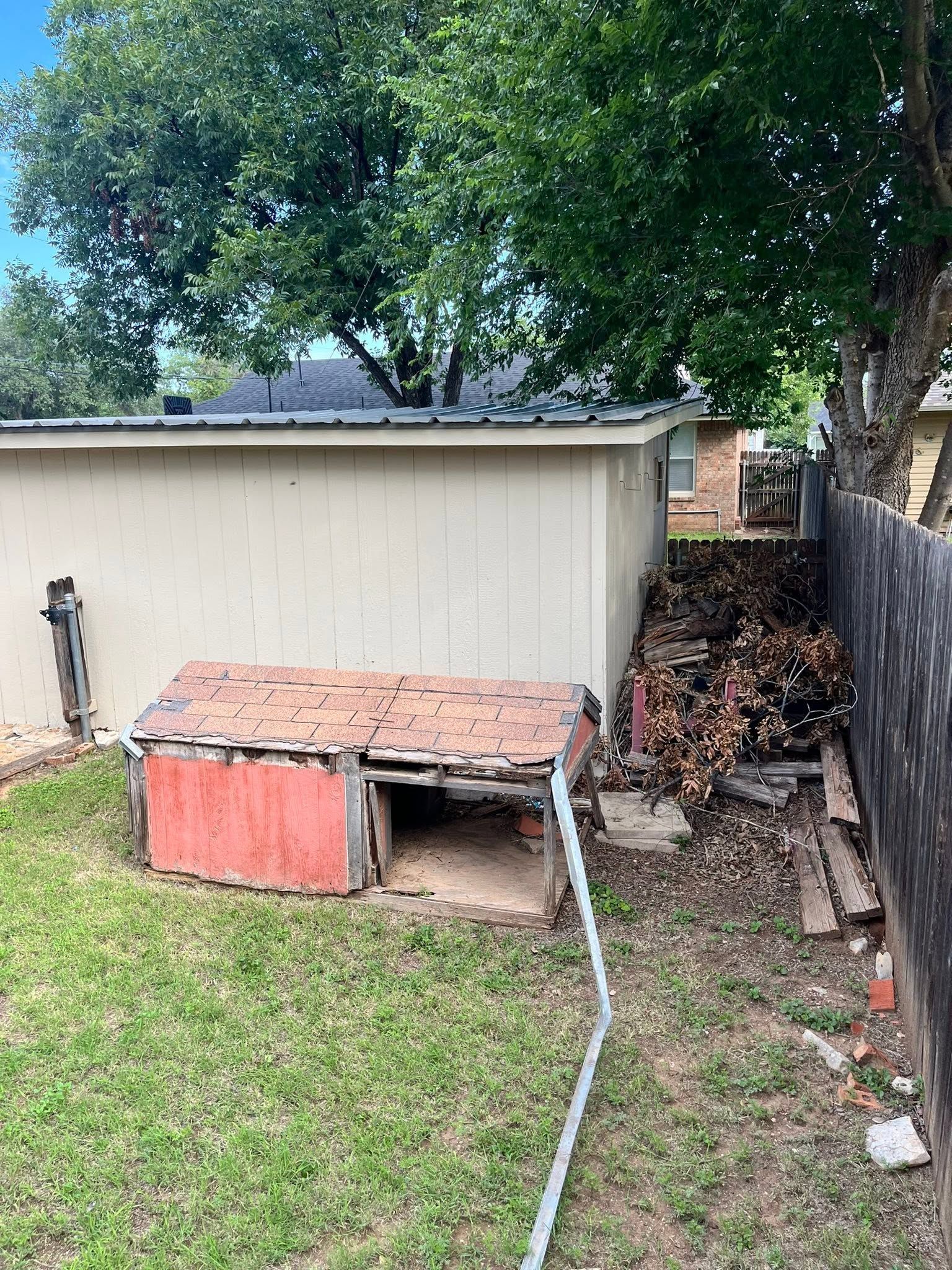 Backyard with beige shed, red doghouse-like structure, tree, and a hose on the ground.
