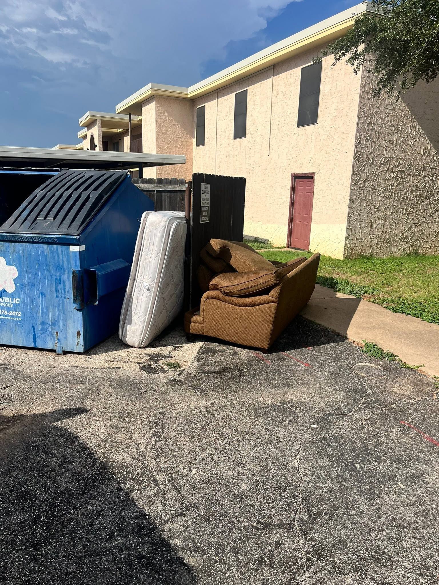 Blue dumpster beside a tan apartment building with stacked furniture on the pavement