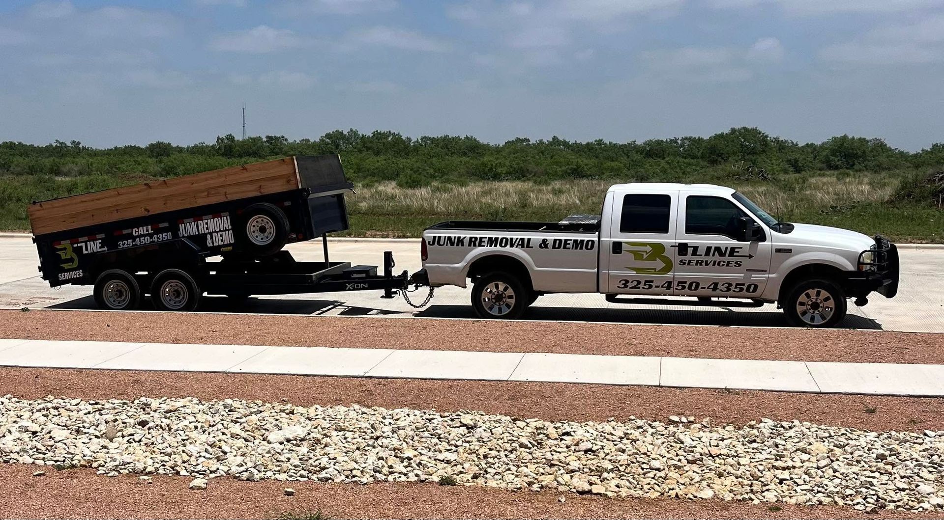 White pickup towing a black dump trailer on a gravel road in a rural area under a cloudy sky