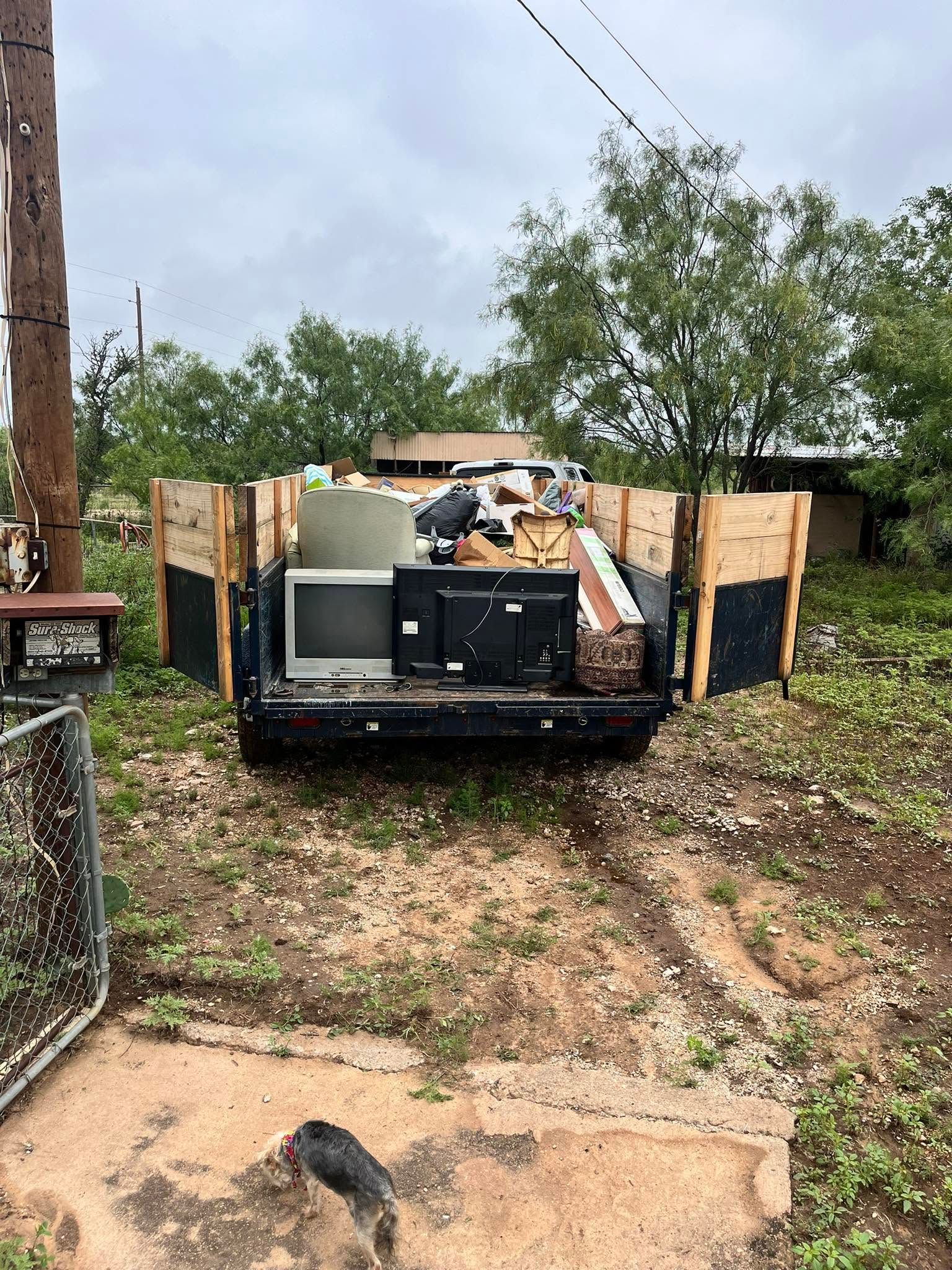 Trash-filled trailer in a muddy yard with discarded furniture and debris behind a chain-link fence