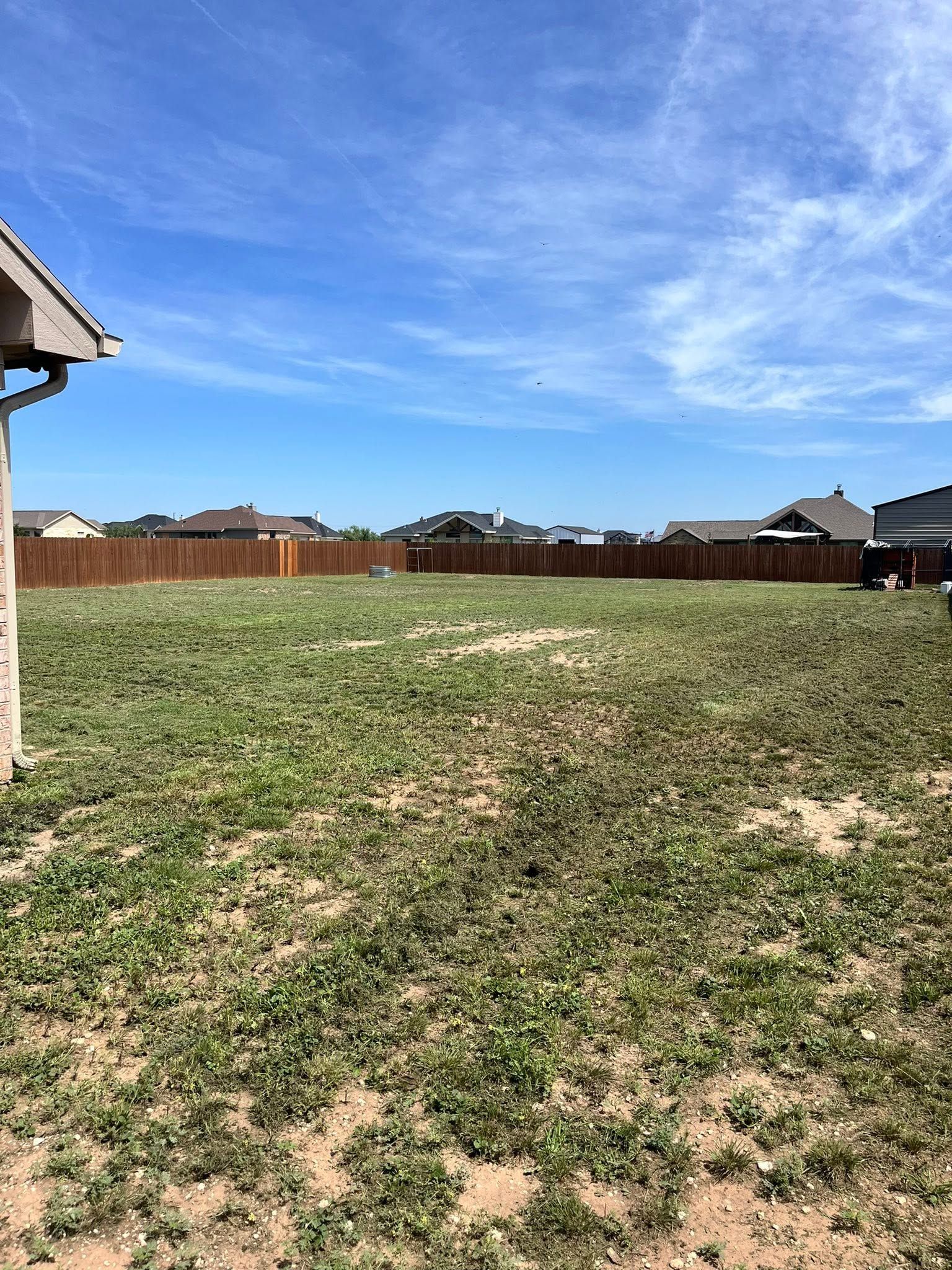 Empty grassy backyard with patchy dry ground, wooden fence, and blue sky with wispy clouds