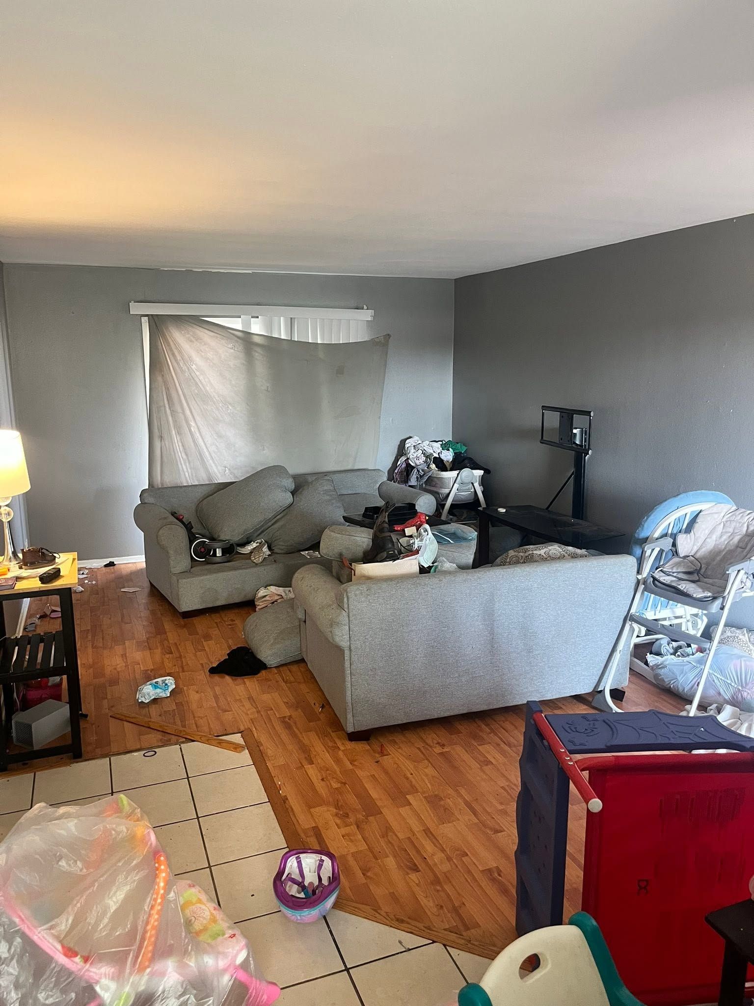 Cluttered living room with gray couch, exercise bike, scattered items, and wood floor throughout