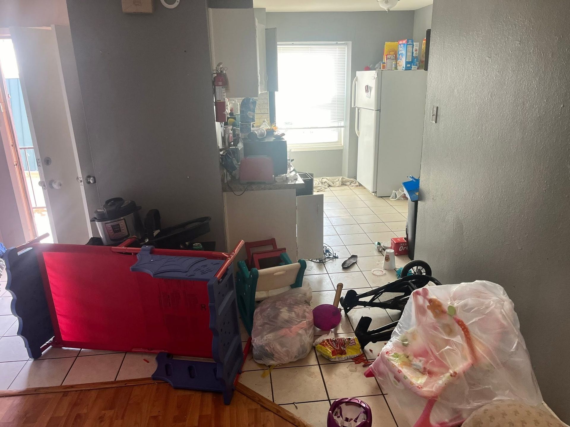Cluttered hallway with scattered items, a red playpen, and a kitchen visible at the end.