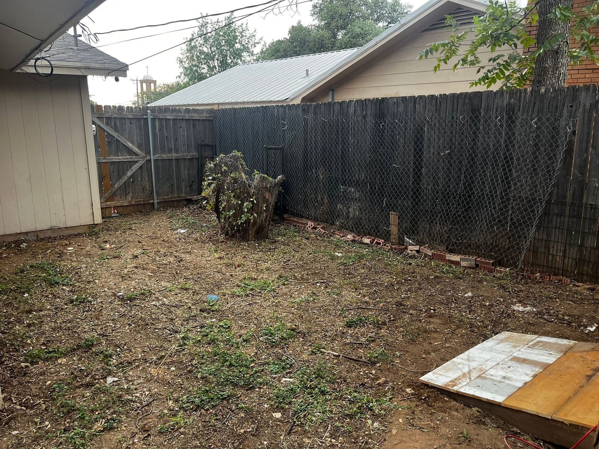 Small backyard with a wooden fence, scattered leaves, and a tarp-covered board in the foreground.
