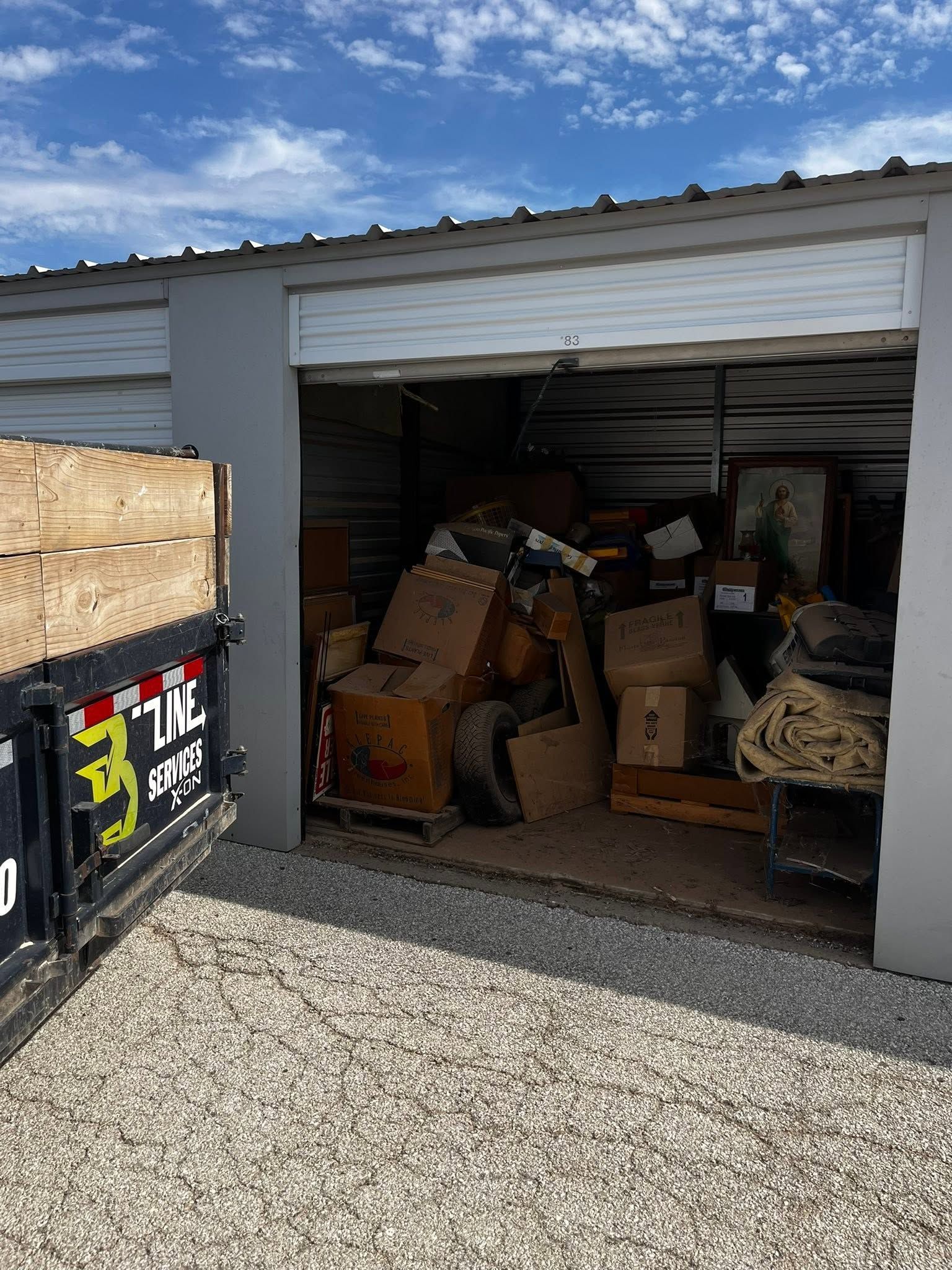 Open garage filled with boxes, furniture, and clutter beside a gravel driveway.