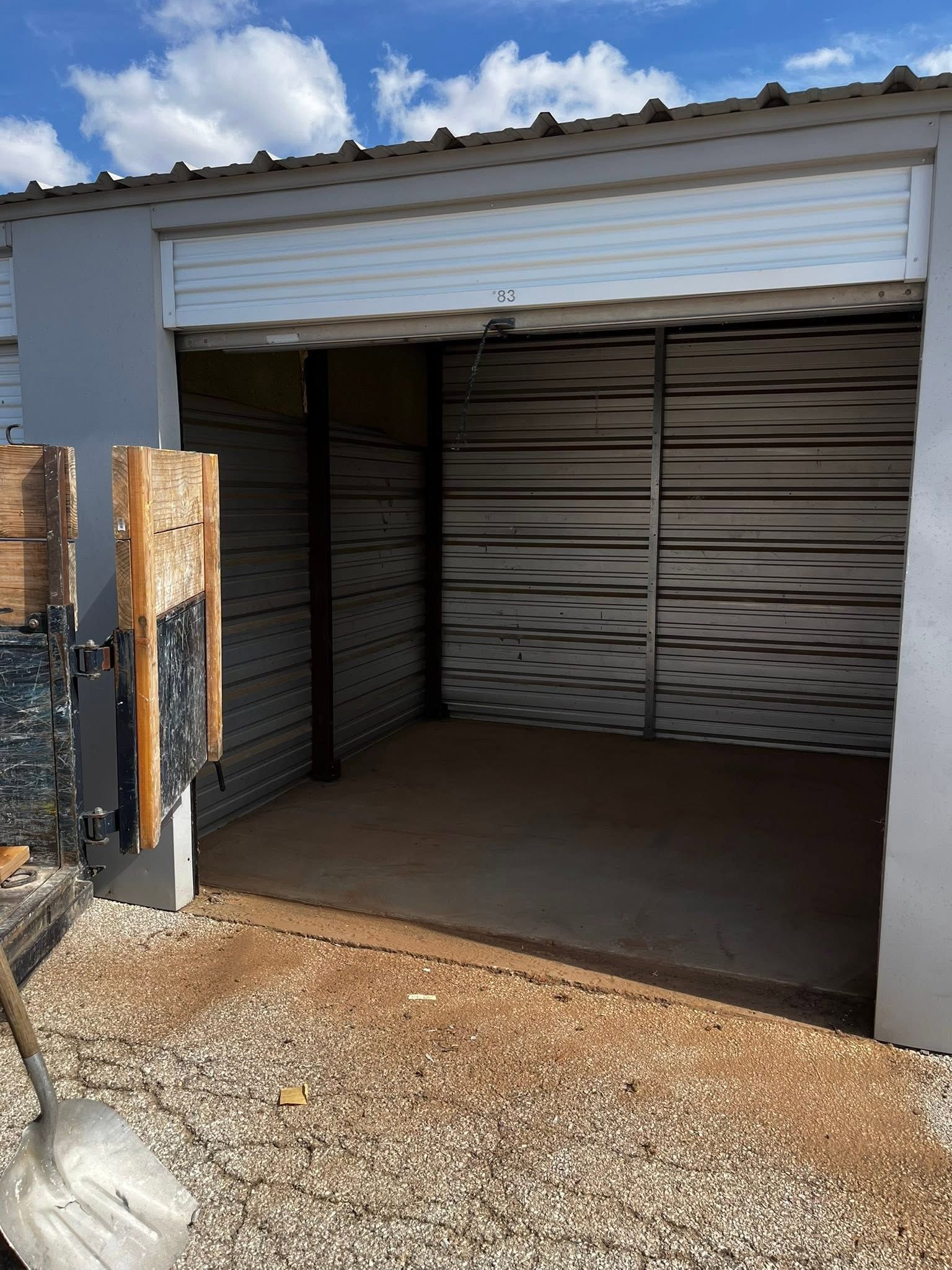 Open storage unit with metal roll-up door and gravel driveway under sunny sky