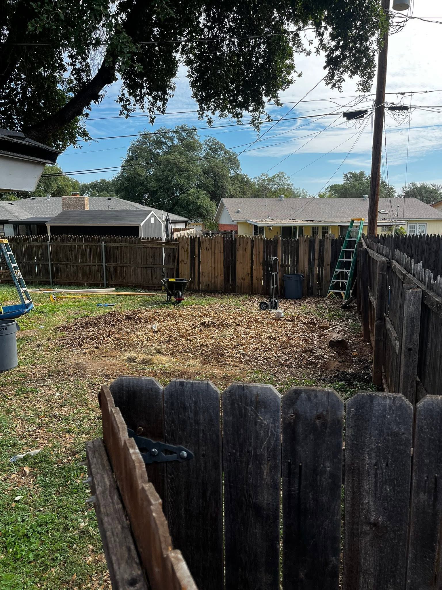 Backyard with wooden fences, fallen leaves, trees, and a cloudy blue sky.