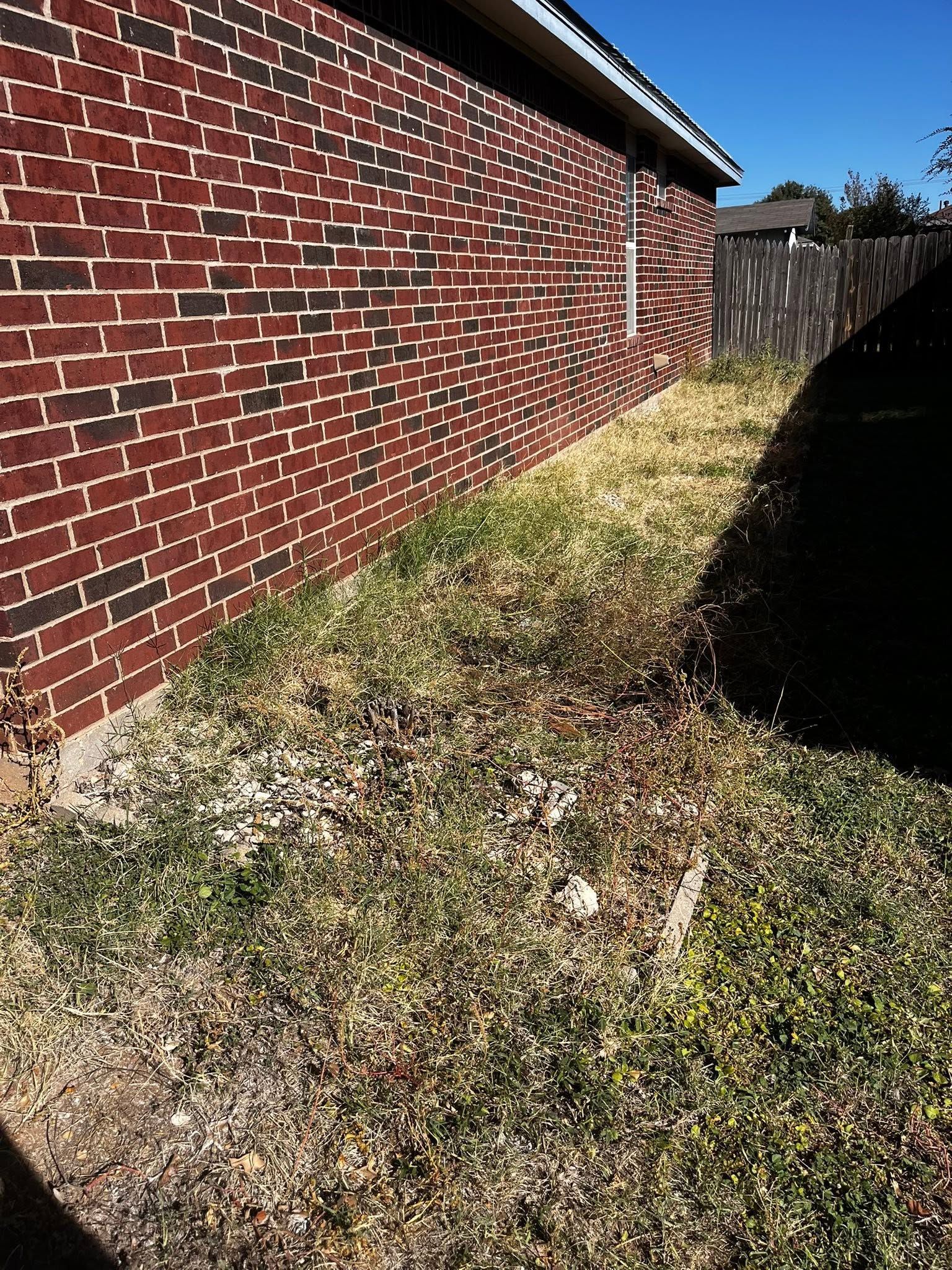 Red brick house wall beside a narrow grassy strip and wooden fence in bright sunlight.