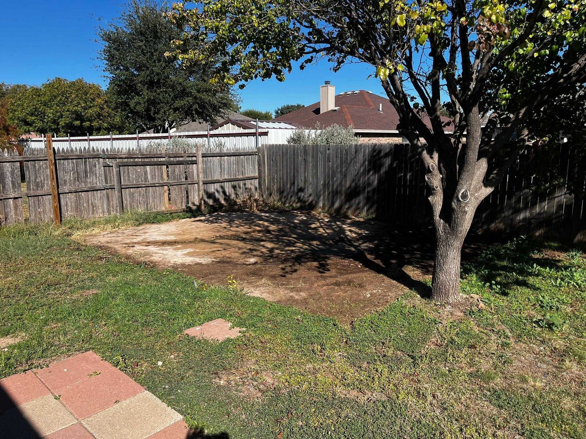 Backyard with a tree, wooden fence, patchy grass, and a bare dirt area under a sunny sky