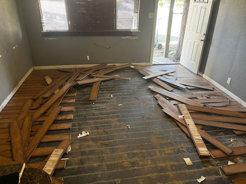 Damaged room floor with torn wooden boards scattered across the room near a window and open doorway