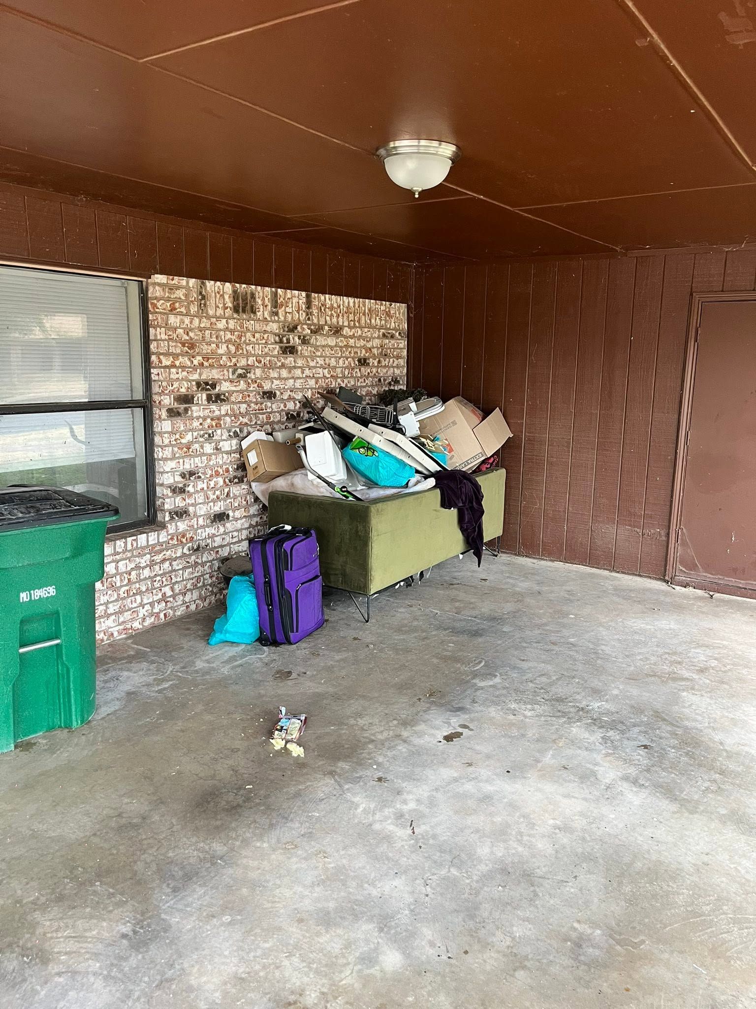 Covered porch with stacked bags and bins beside a brick wall and green trash can.