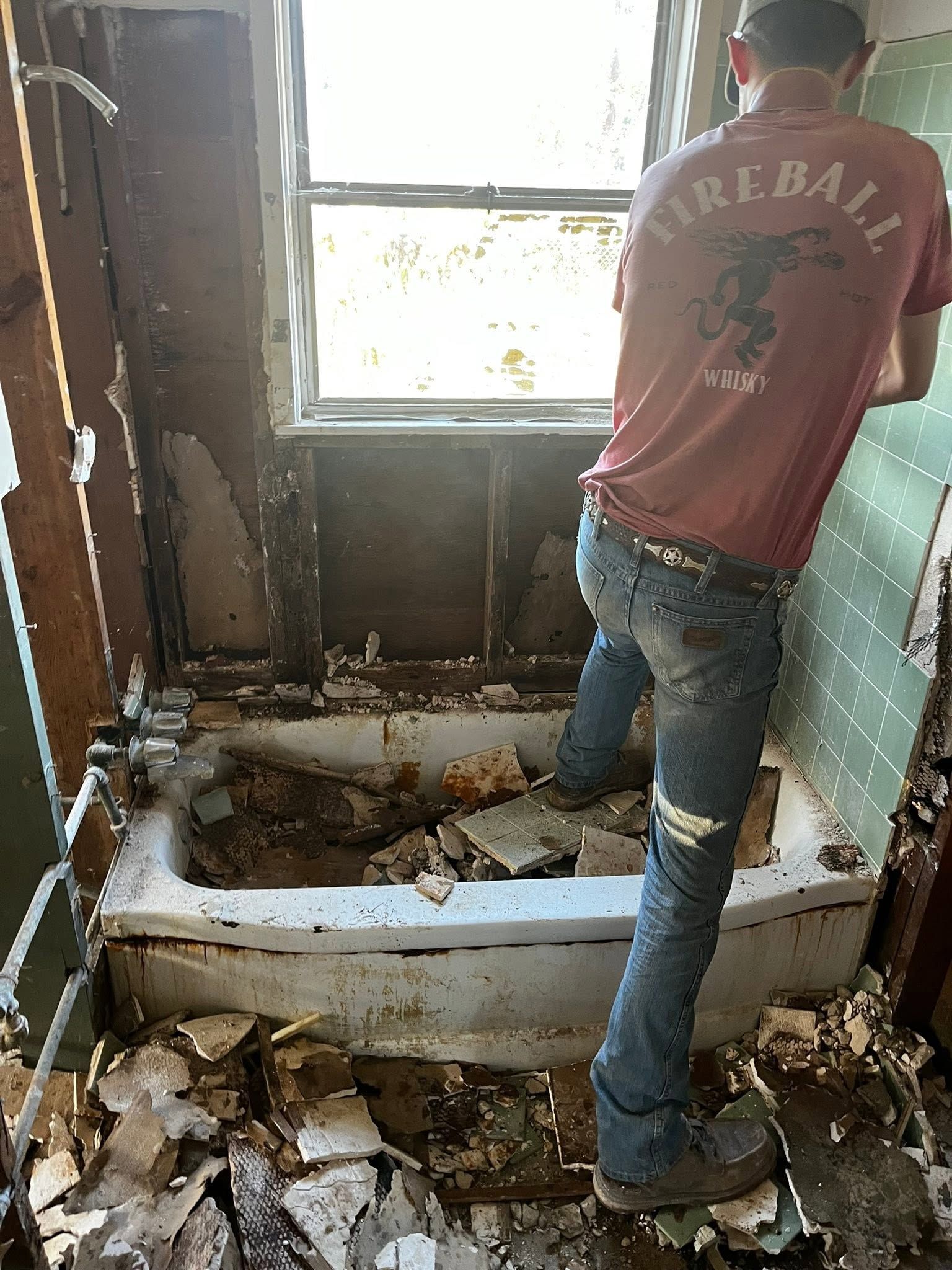 Person standing in a torn-up bathroom with a broken bathtub, debris on the floor, and peeling walls.