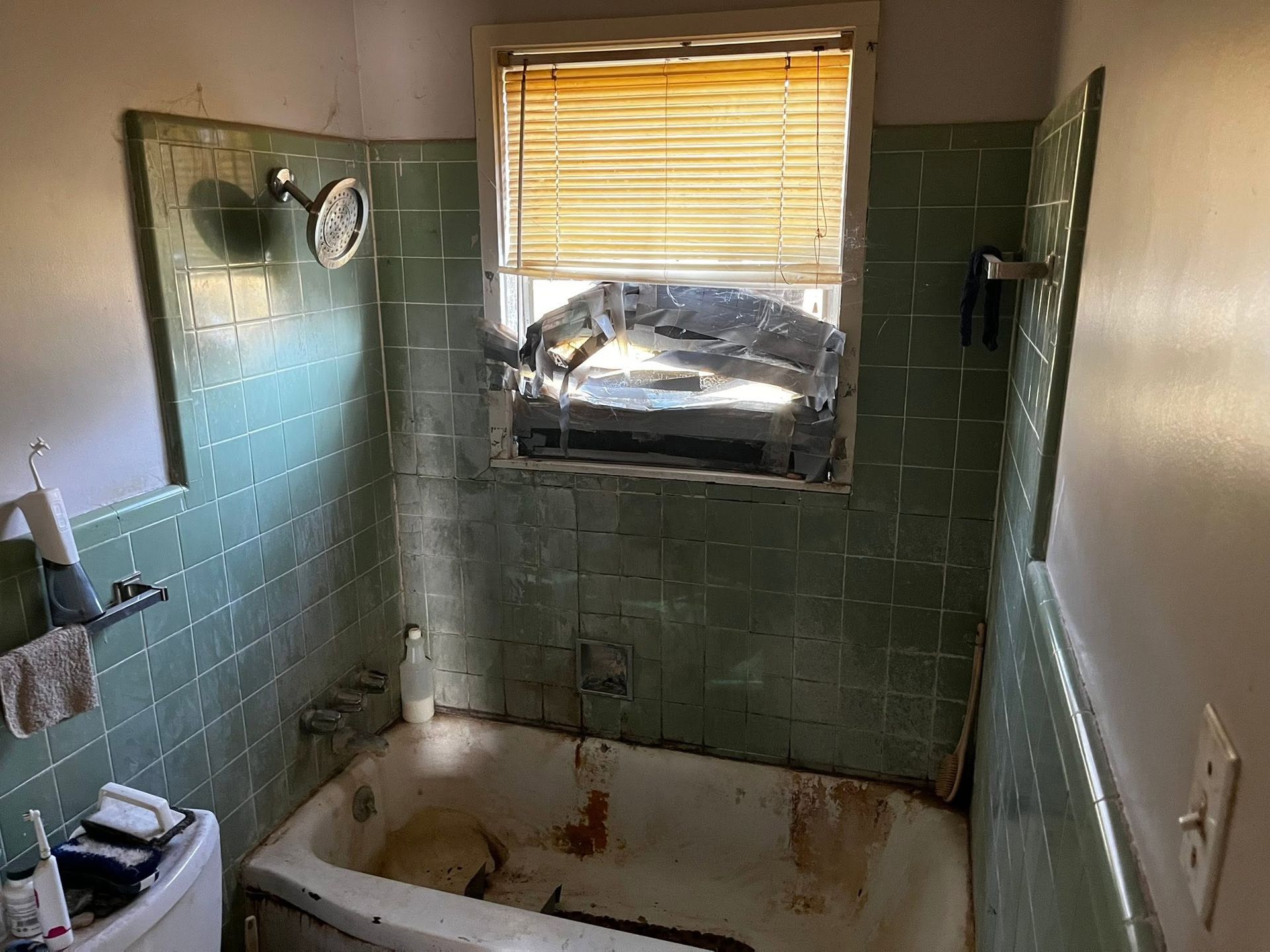 Small, grimy bathroom with green tile, stained bathtub, and a window with blinds above the tub