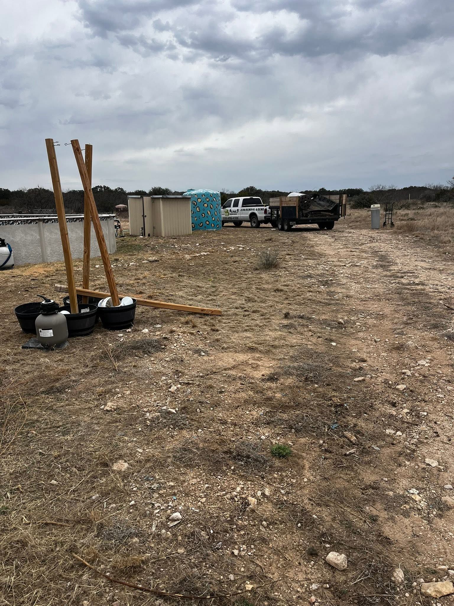 Rocky open lot with planted saplings in pots, fencing, and a truck under a cloudy sky